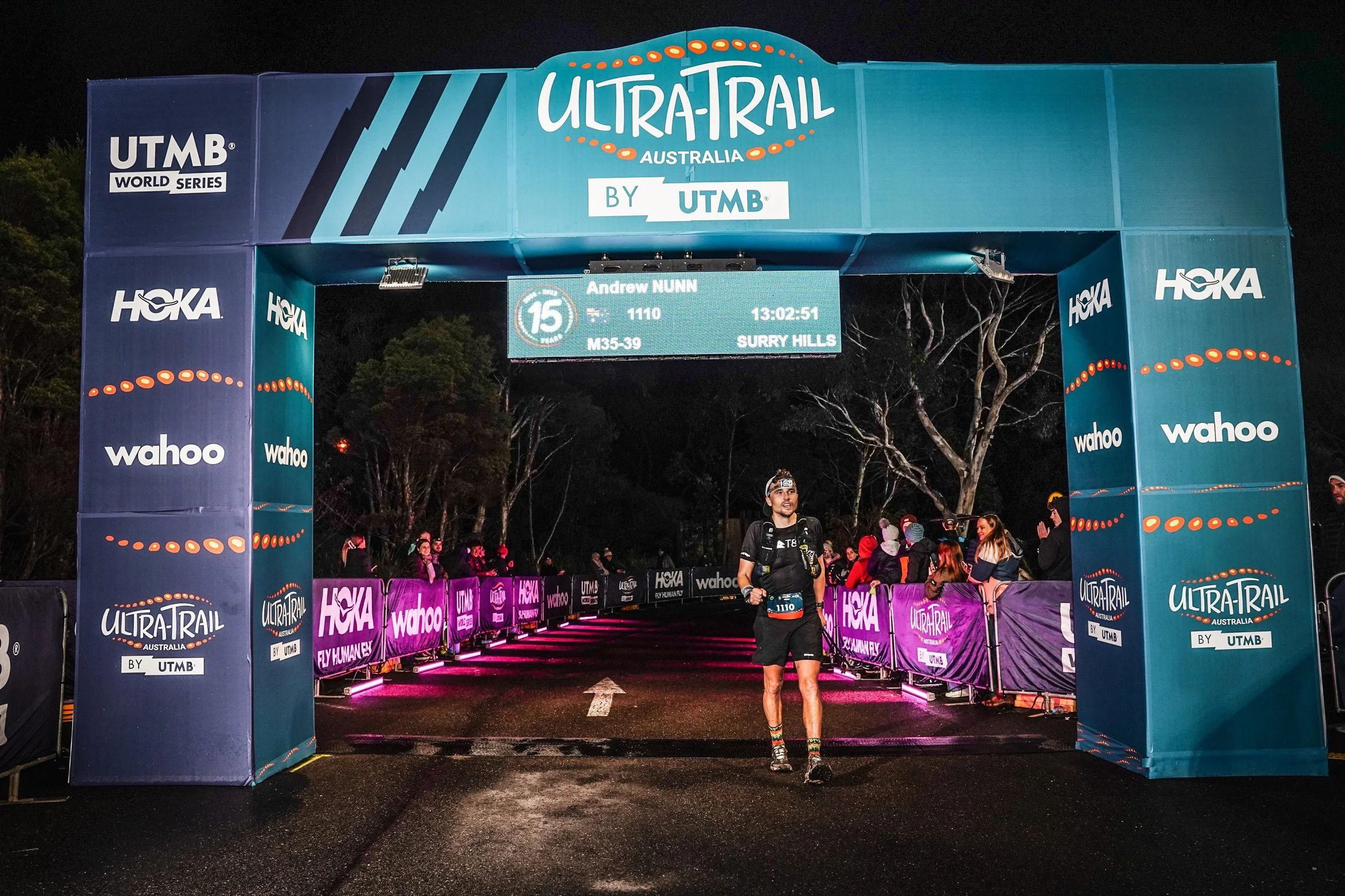 A male runner crosses the finish line at the Ultra-Trail Australia race during night time, with spectators behind barriers watching. The finish line arch displays sponsor logos and event details.