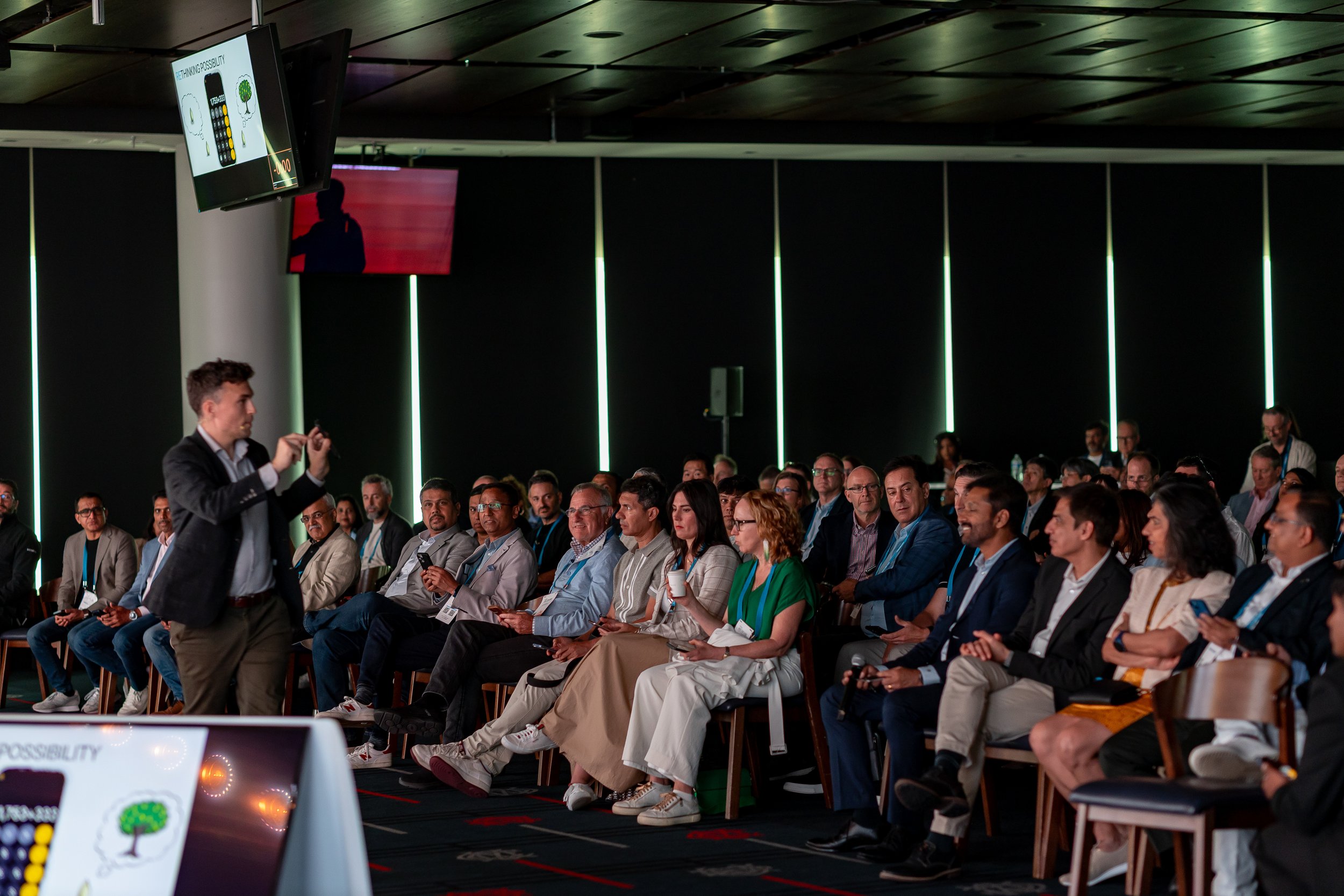 A speaker presenting to an audience at a conference or seminar in a dark room with vertical green light strips on the walls. The audience is seated and attentively listening.