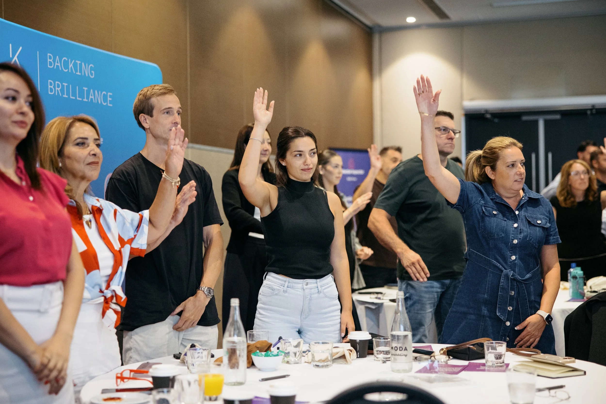 Group of diverse people raising their hands in a conference room with a blue sign that says 'Backing Brilliance'.