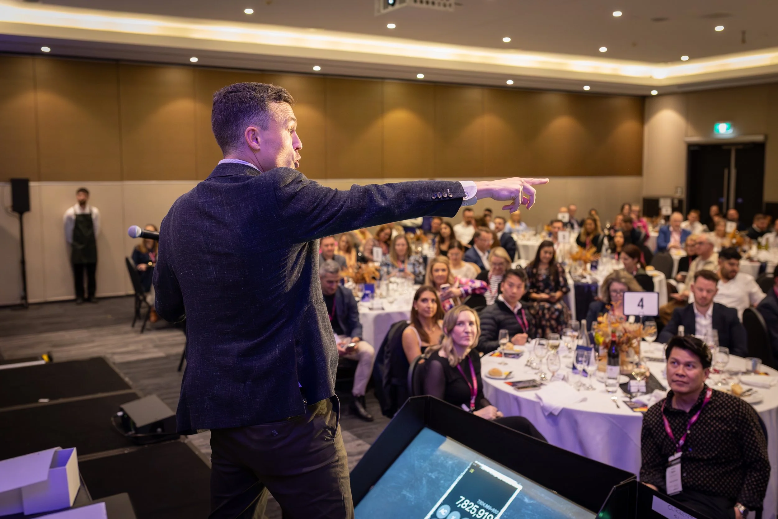A man in a suit speaking or presenting to an audience in a conference or banquet hall filled with seated attendees.