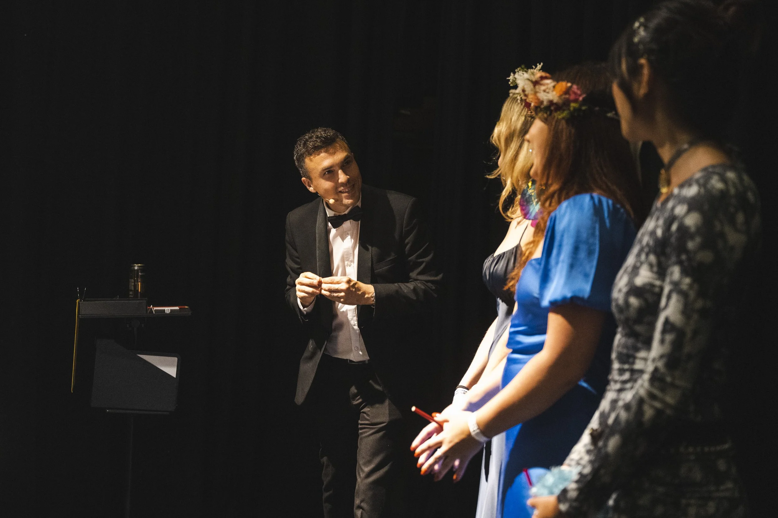 A man in a tuxedo is speaking to a group of four women on a stage with a black background, possibly during a performance or presentation.