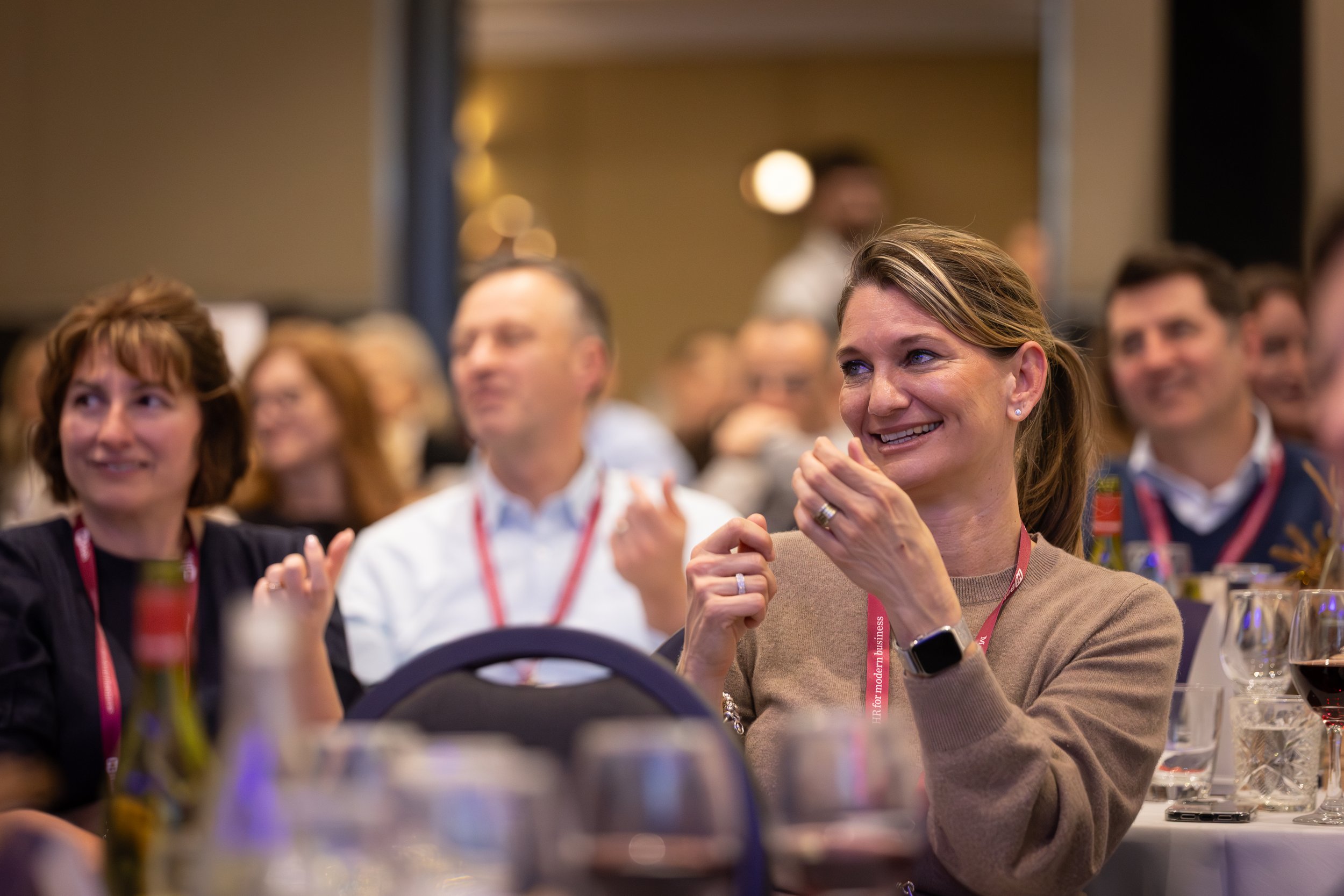 People attending a conference or seminar, sitting at tables with drinks, listening and smiling.