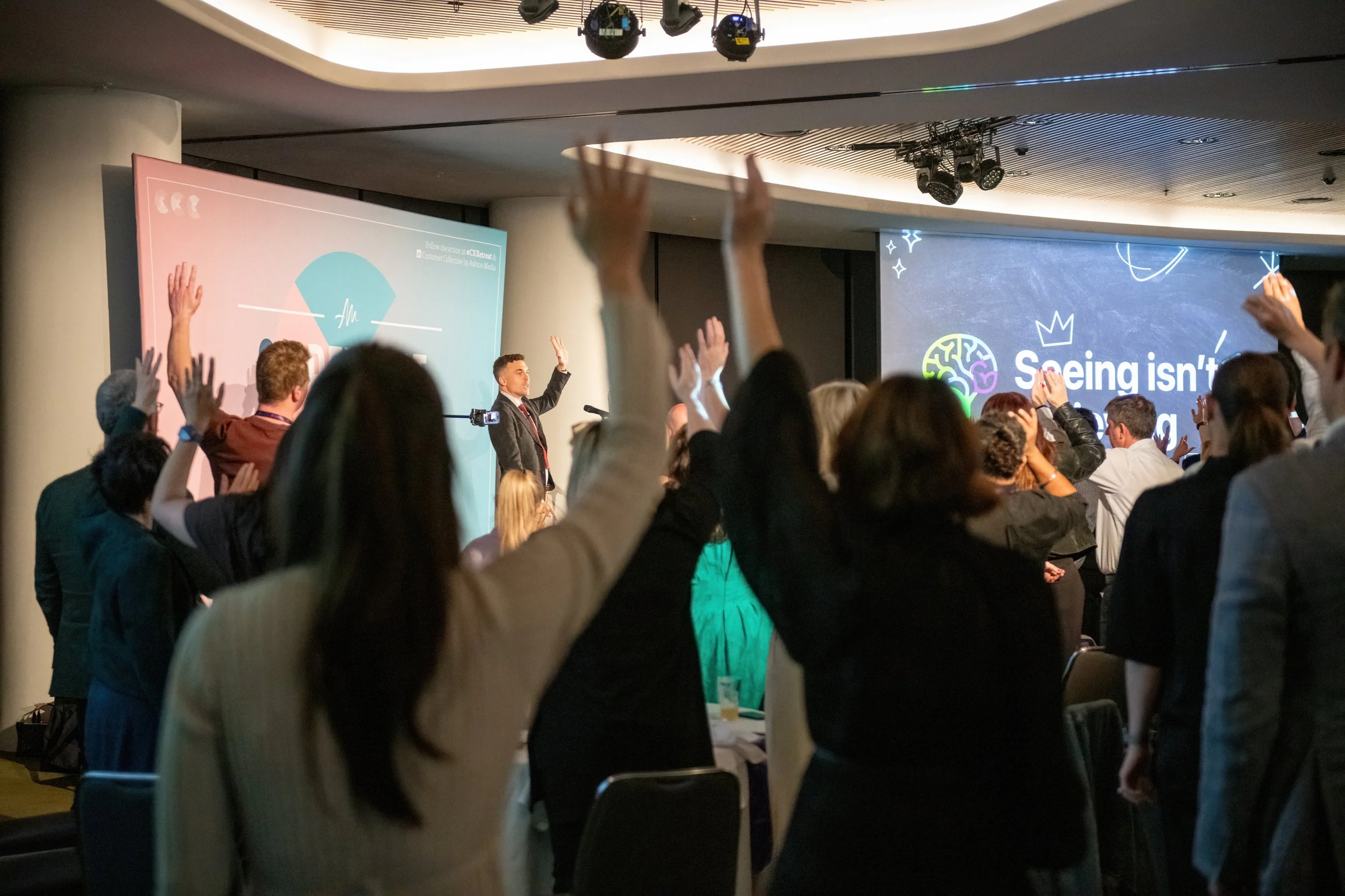 A conference room filled with people raising their hands while a speaker stands near a large screen.