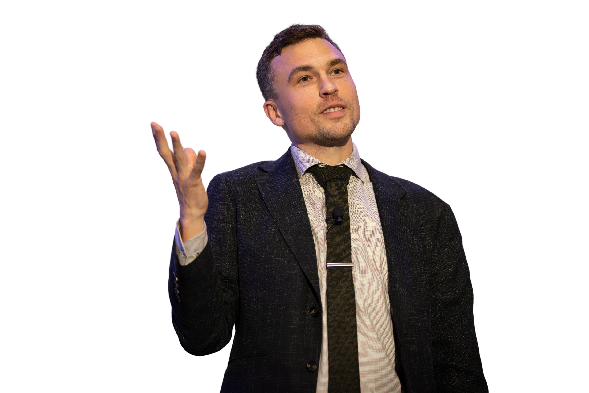 A man in a suit giving a presentation, gesturing with his right hand, with a blurred background.