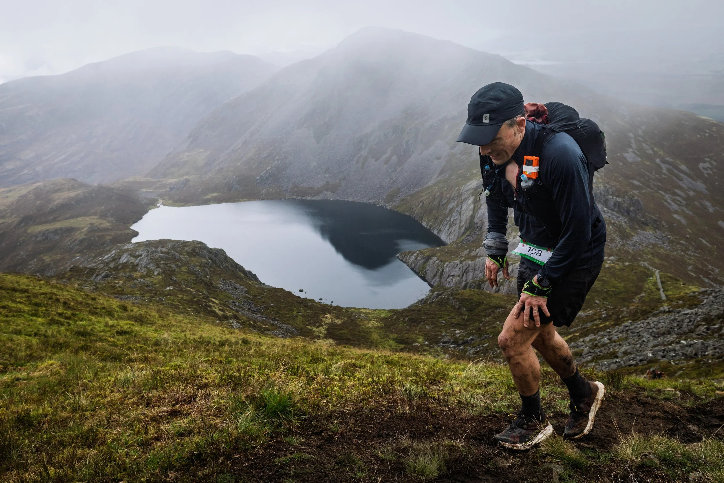 A man running on a muddy trail in a mountainous area with a lake in the background, wearing sports gear and a backpack.