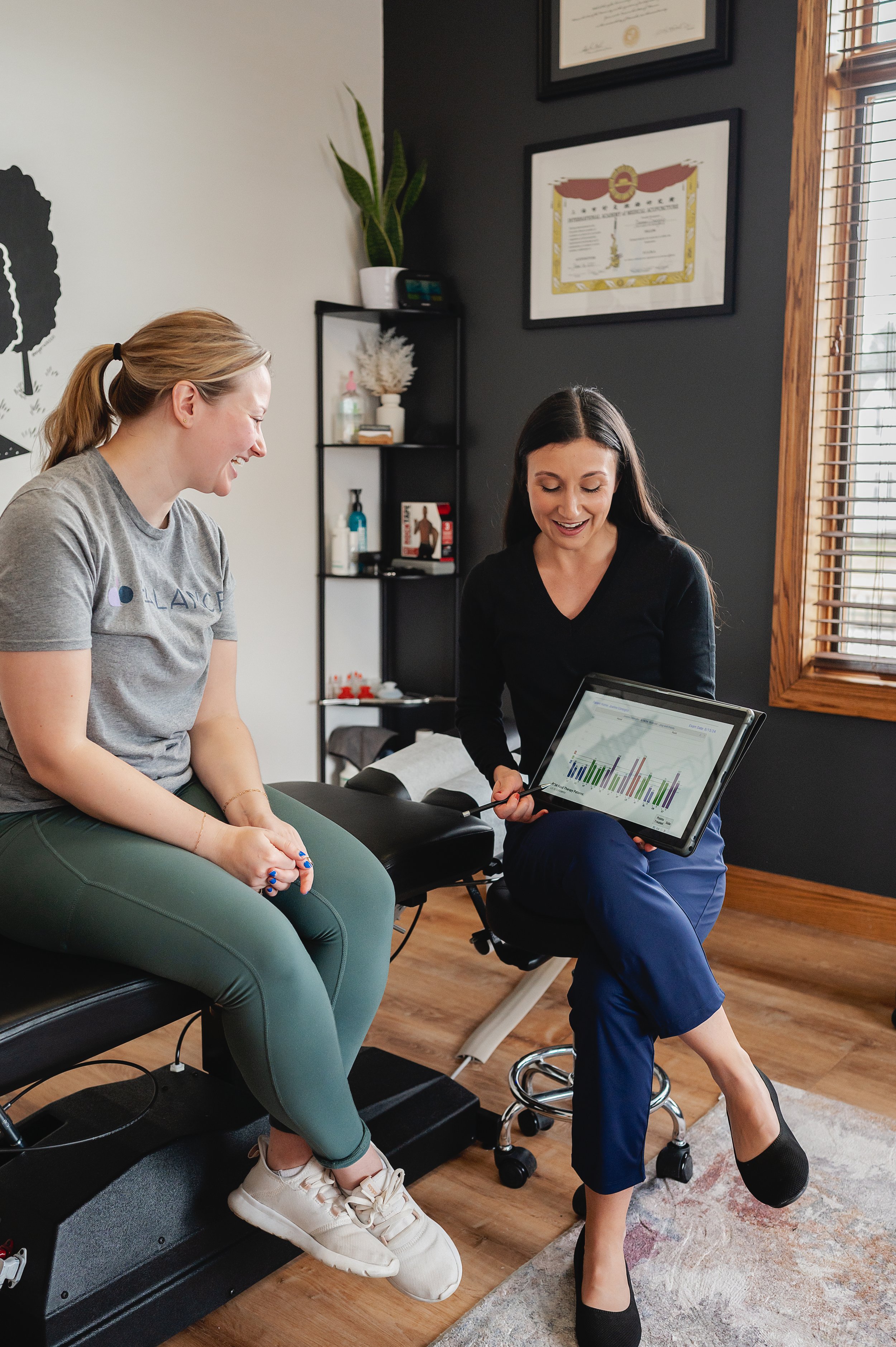 A woman with long dark hair sitting on a medical examination table, smiling and holding a tablet showing a colorful bar graph, while a woman with blonde hair in a ponytail sitting on the edge of the table laughs. The room has a black and white wall, framed certificates, a window with blinds, and a shelf with small decor and medical supplies.