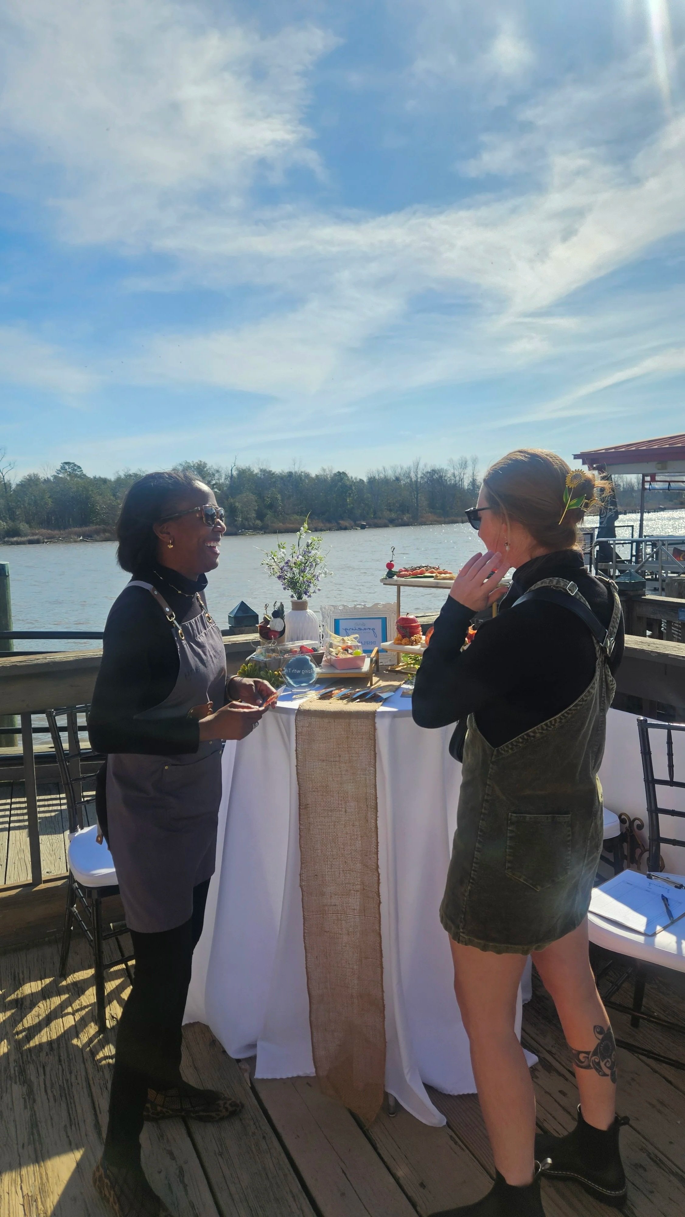 Two women are having a conversation outdoors by a river, with a small table decorated with food and flowers between them. The woman on the left is wearing a black apron and sunglasses, and the woman on the right has hair in a bun with flowers The sky is partly cloudy and the scene is bright and sunny.