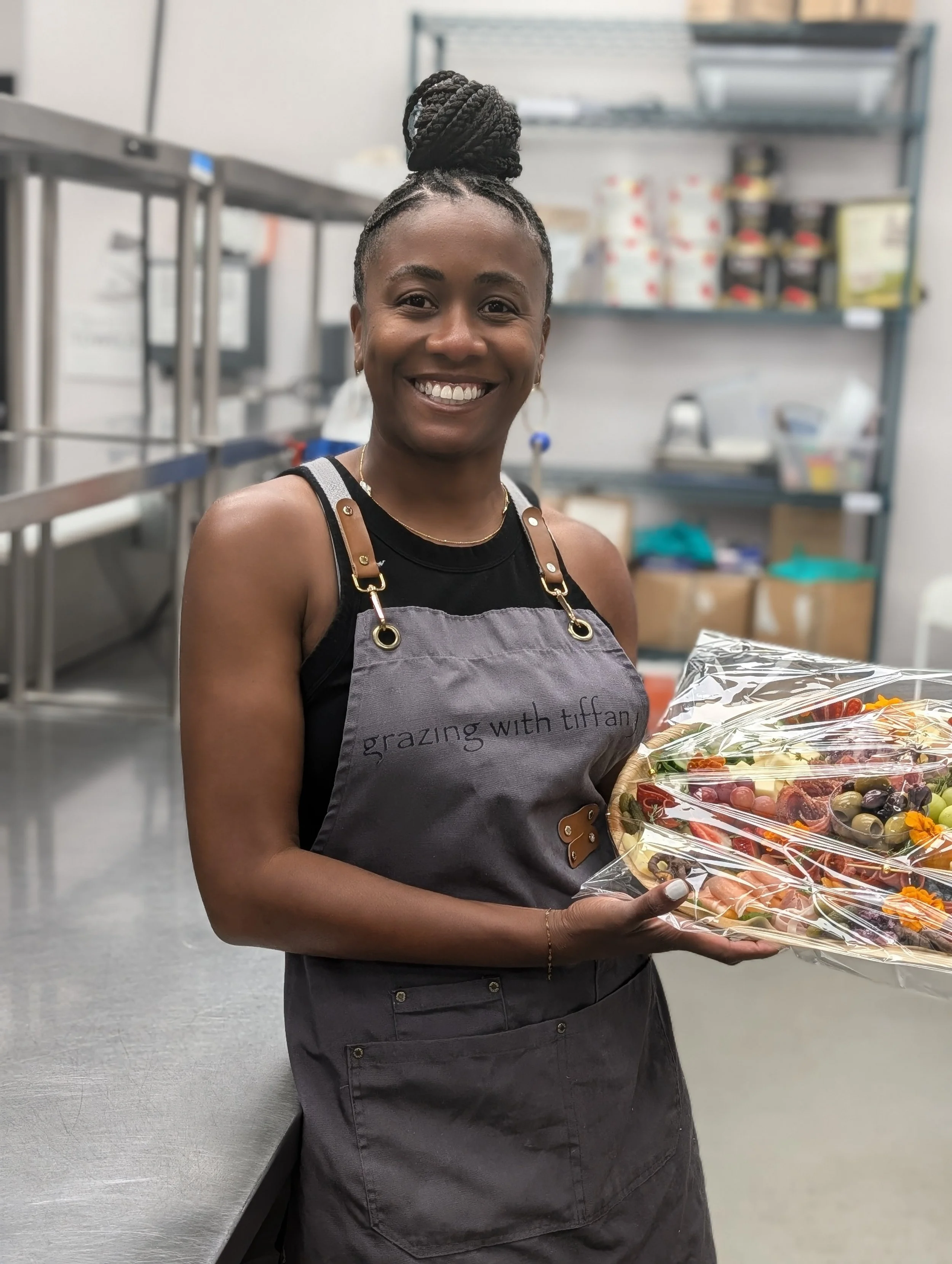 Smiling woman wearing a gray apron, holding a charcuterie board in a kitchen