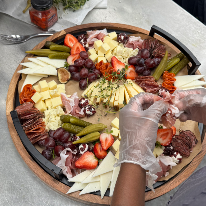 Person arranging various cheeses, strawberries, grapes, pickles, and cured meats on a large wooden serving platter for a charcuterie board, wearing gloves.