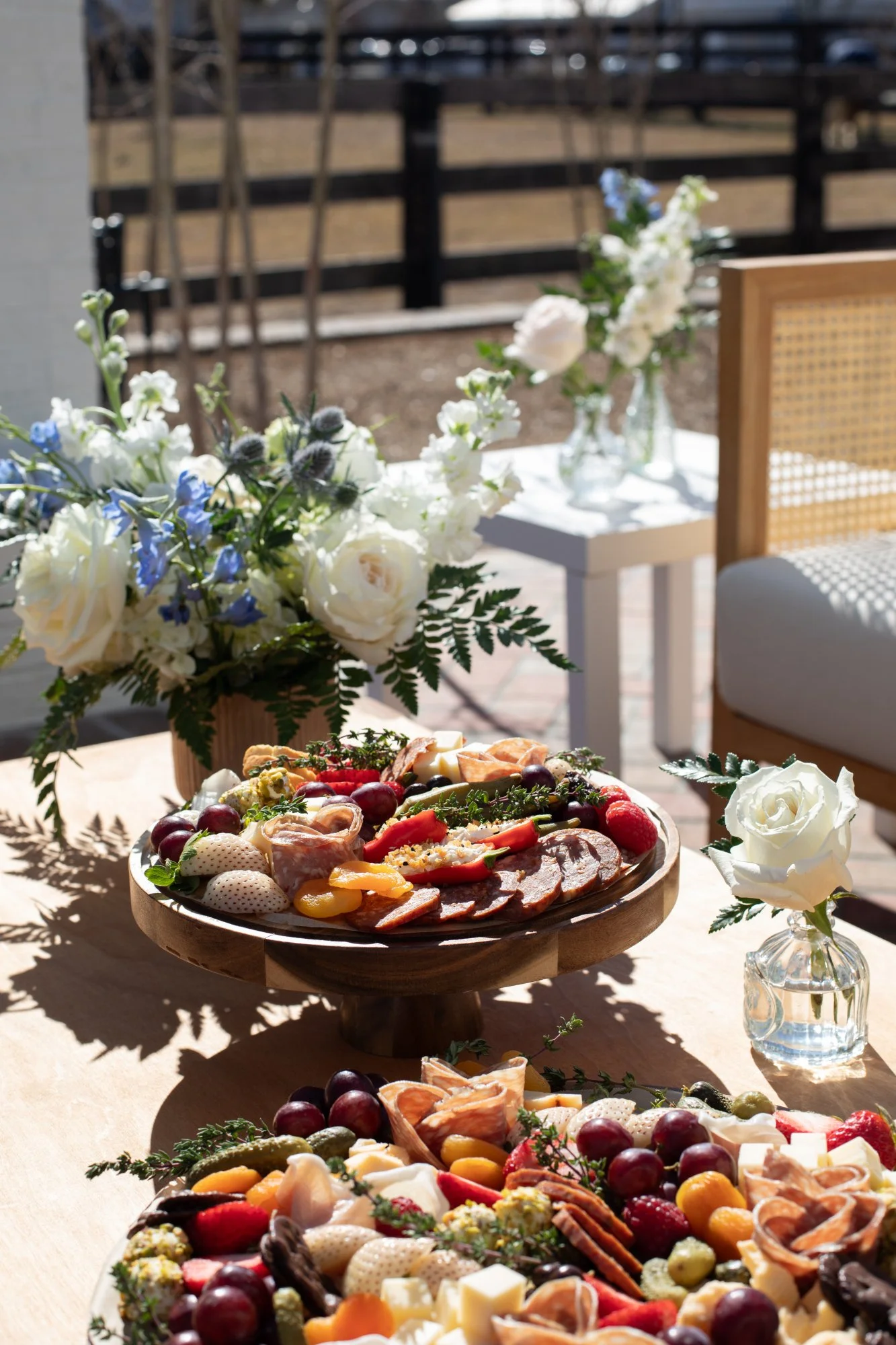 Table with assorted charcuterie, fresh fruits, and flowers in vases at an outdoor event.