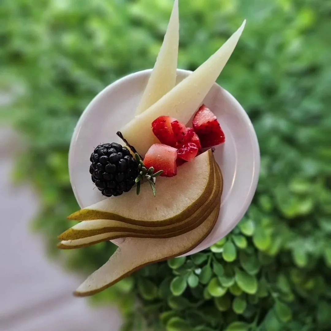 A bowl of fresh sliced pears garnished with sliced strawberries, a blackberry, and white chocolate shavings, set against a blurred green leafy background.