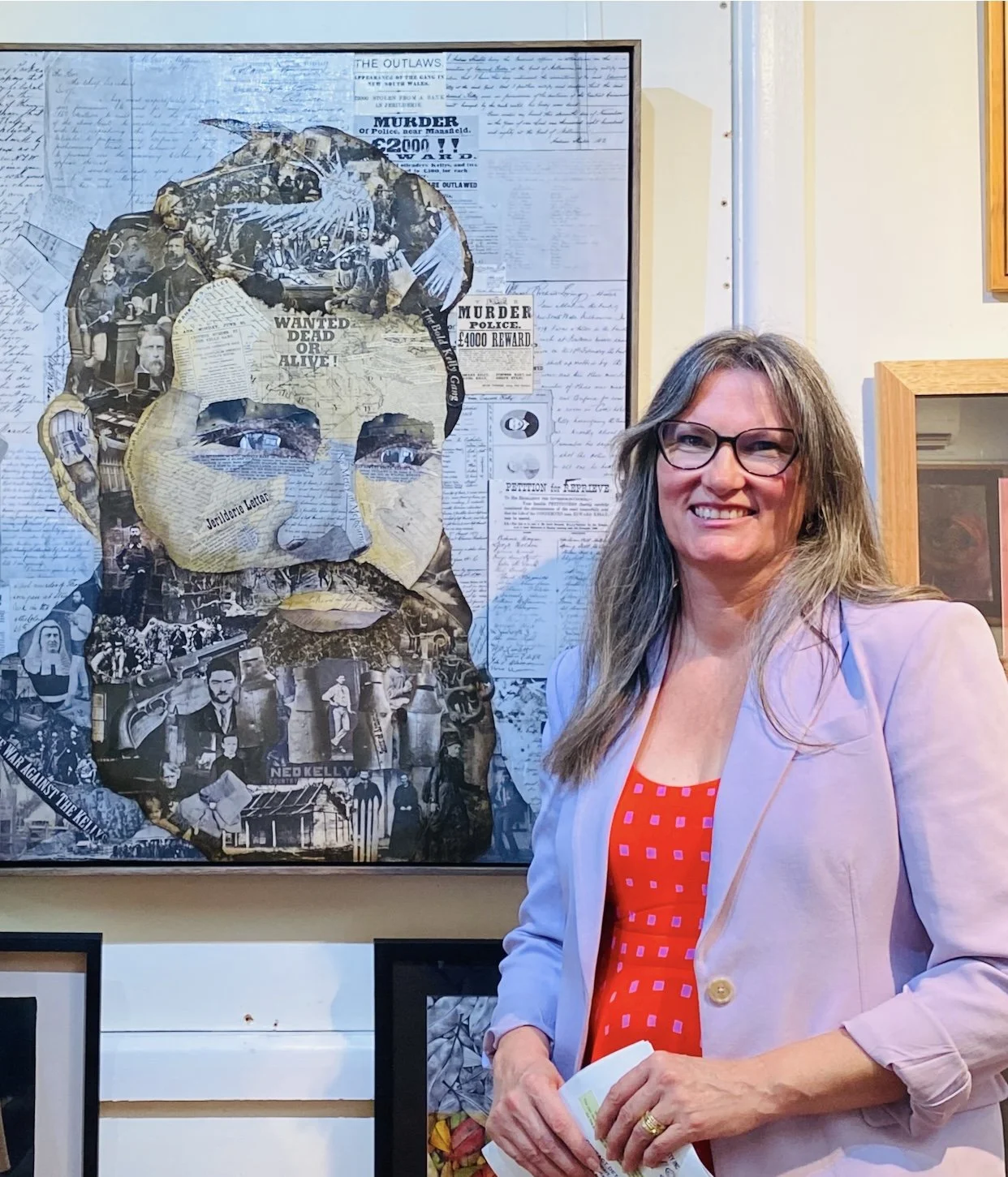 A woman with glasses and long hair smiling, standing next to a collage portrait of a man's face made from newspaper clippings and photographs, displayed on a wall.