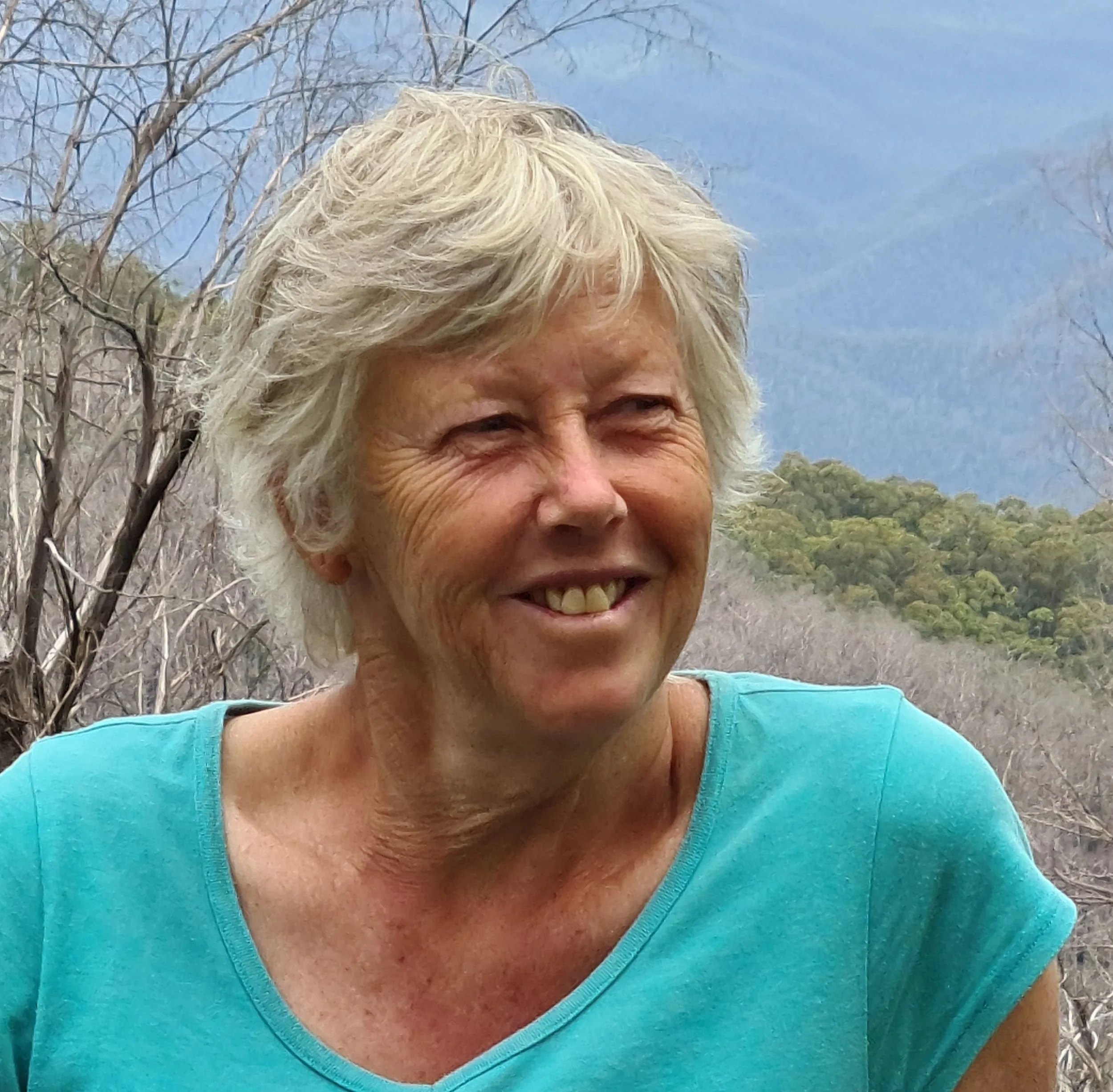 A woman with curly hair wearing a blue shirt, standing in front of a bookshelf, holding a painting of a dog's face.