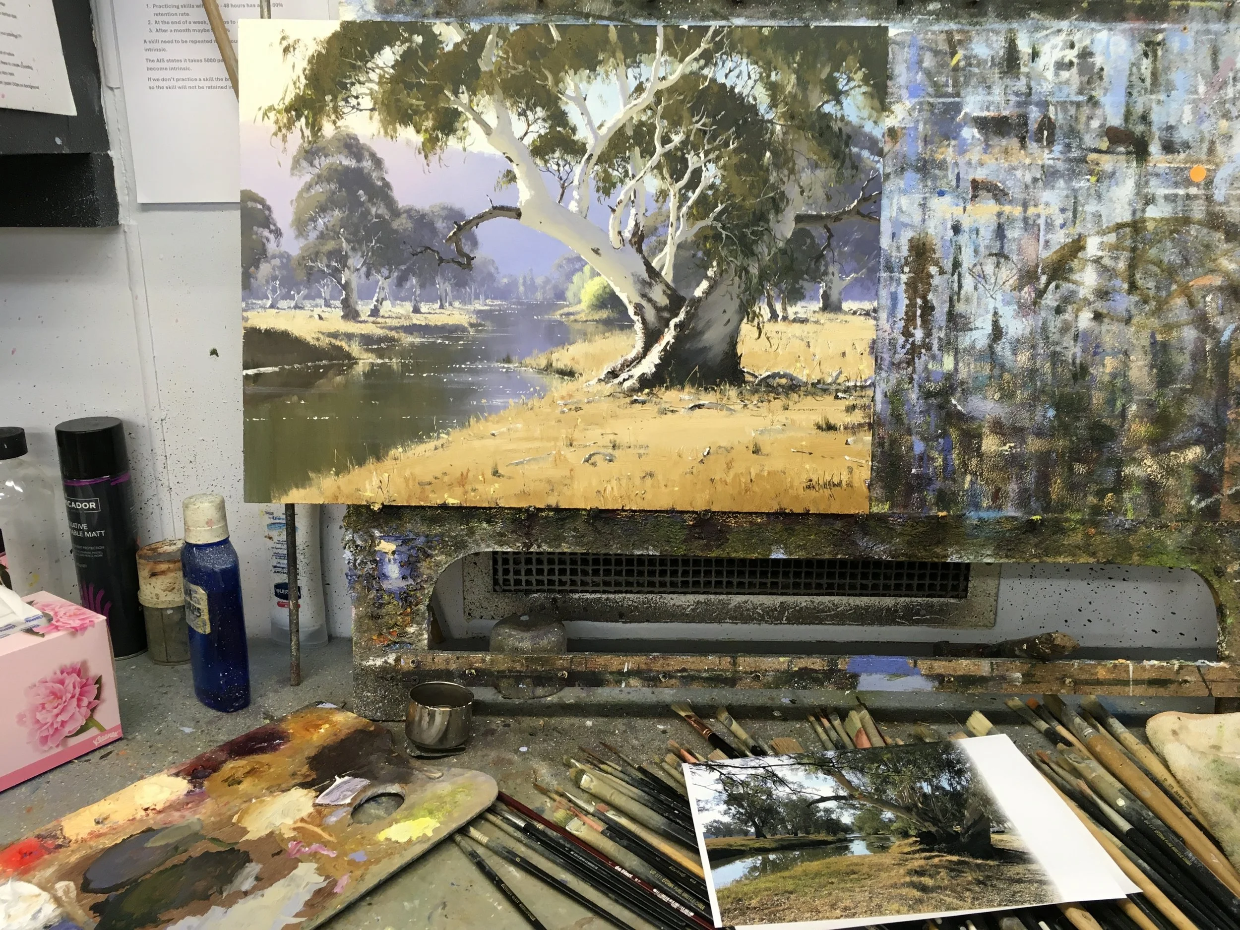 Painting of a landscape with trees and a river, set on an art studio table with brushes, paints, and reference photo.