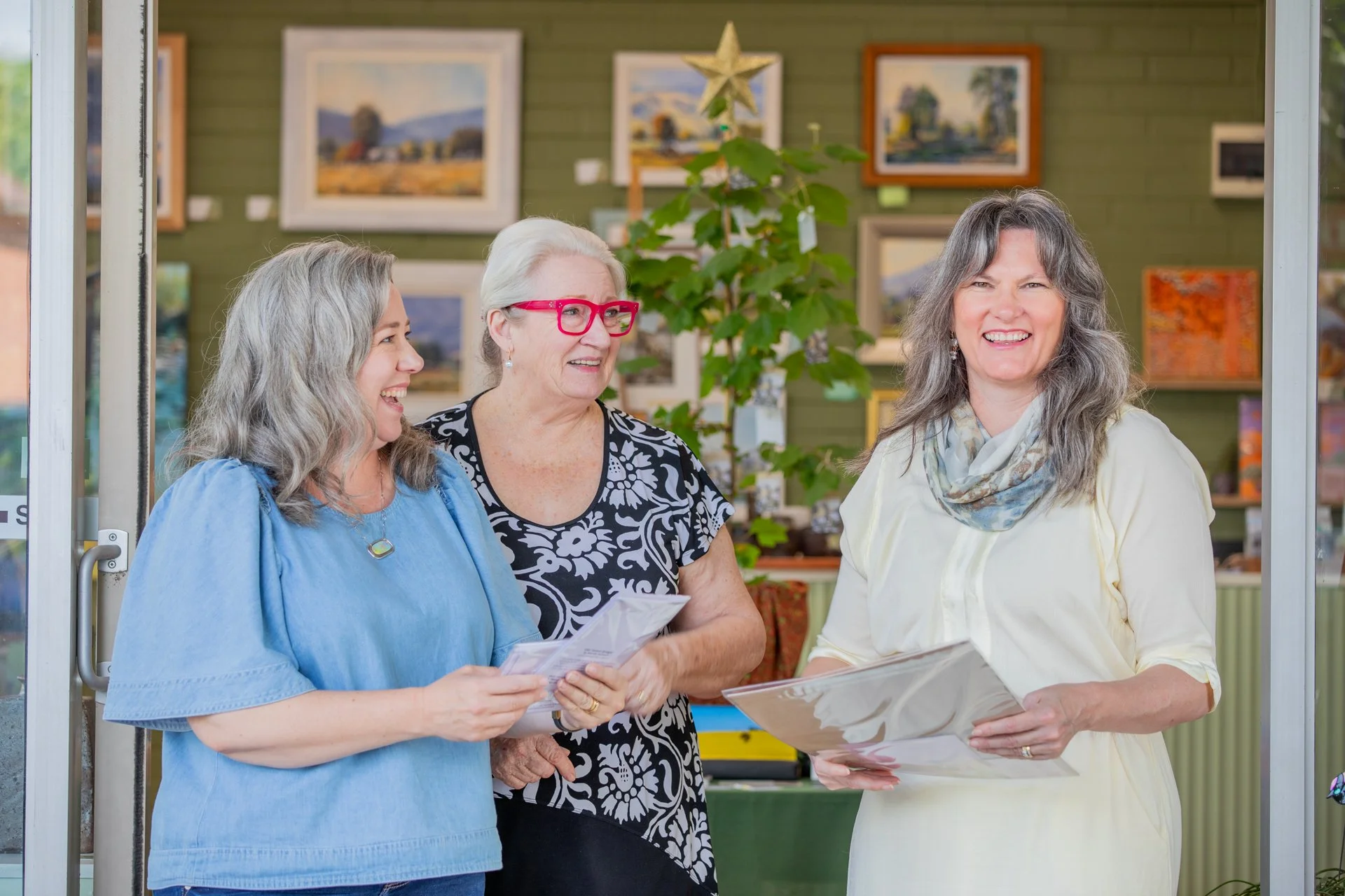 Three women standing and talking in an art gallery, smiling and holding artworks.