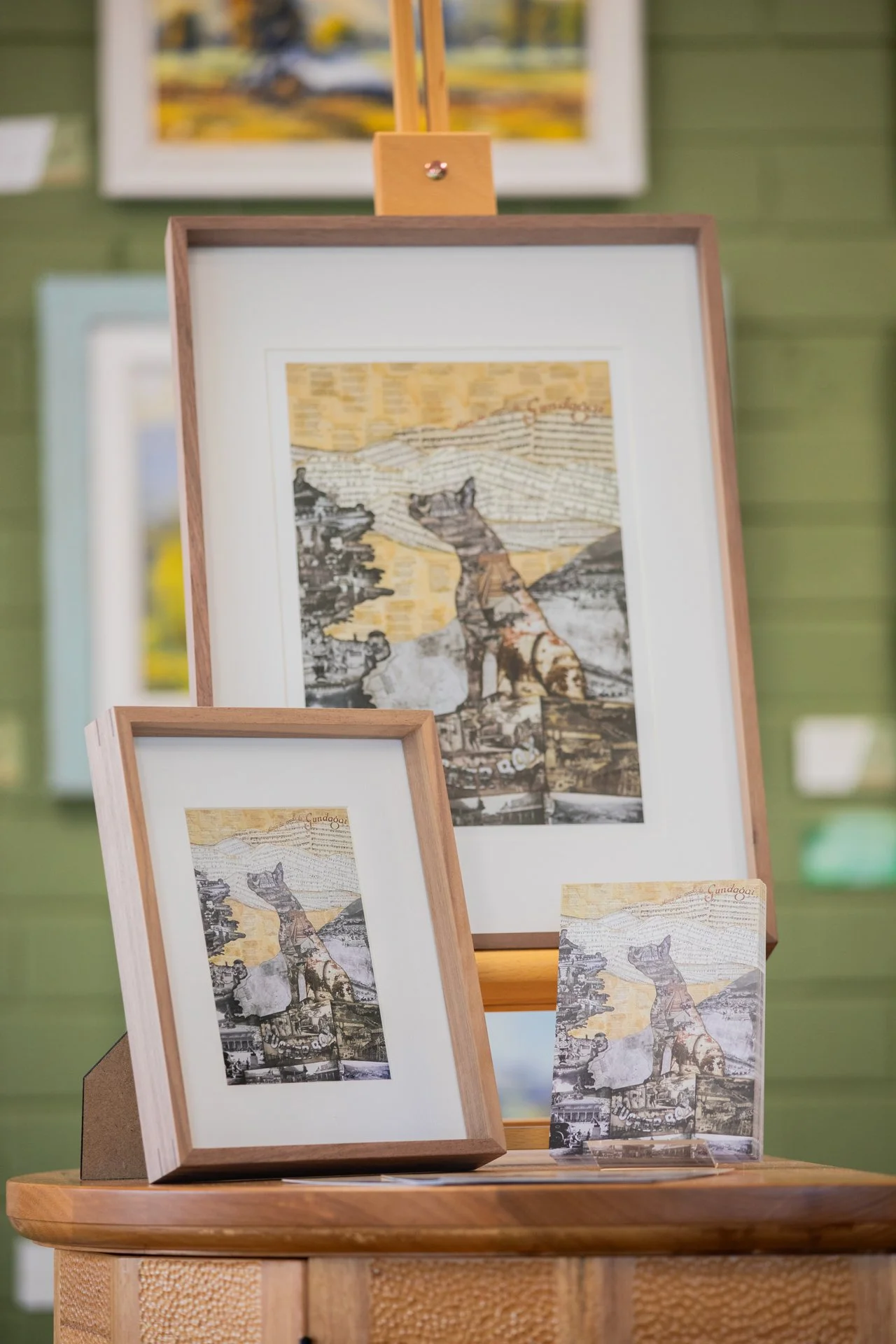 Multi-layered collage artwork of a dog sitting with a landscape background, framed and displayed on a small table, with similar artwork on a larger easel in the background.
