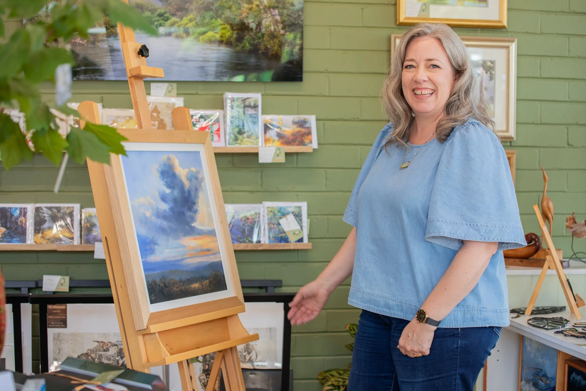 A woman with gray hair smiling inside an art gallery, standing next to an easel with a landscape painting of a cloudy sky and sunset. The gallery has green walls with framed artwork and prints displayed.