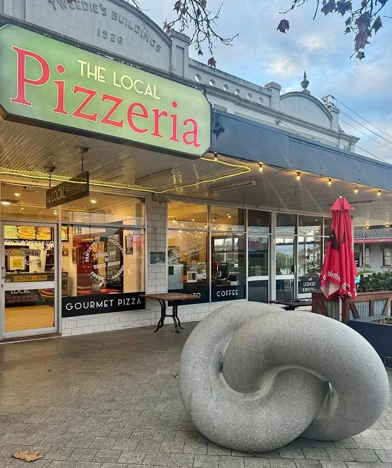 Exterior of a local pizzeria with a large sign reading "The Local Pizzeria," glass windows, and a patio featuring a large, abstract granite sculpture and a red umbrella.