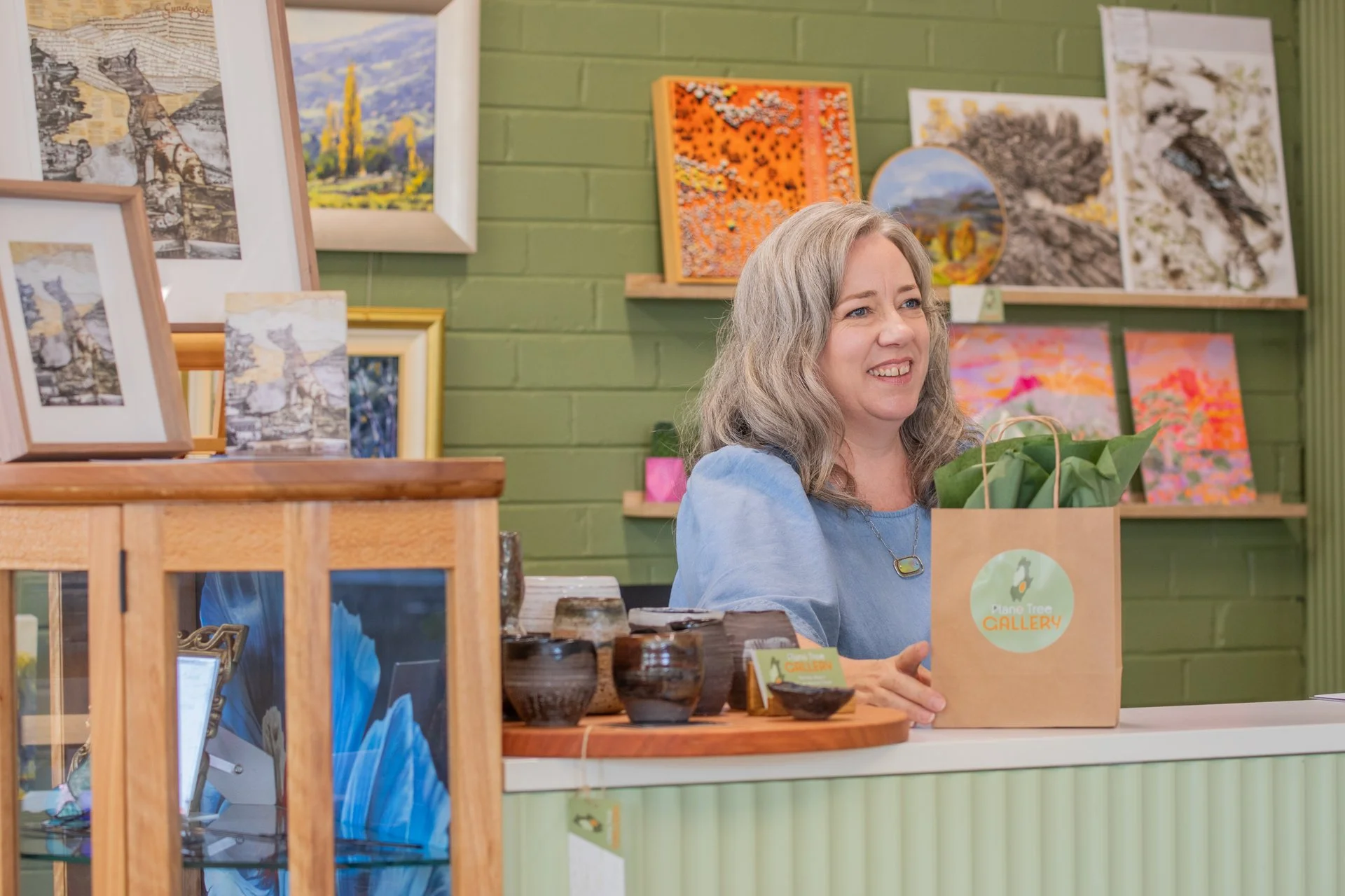 A woman sitting at a counter in an art gallery, smiling, with paintings displayed on a green brick wall behind her, and a paper shopping bag with a green logo in front of her.