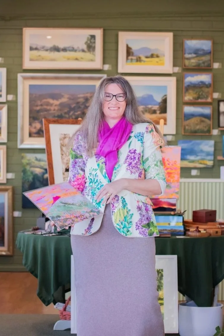 A woman with glasses and long gray hair standing in front of framed landscape paintings at an art gallery, holding a colorful painting, wearing a floral blazer and pink scarf.