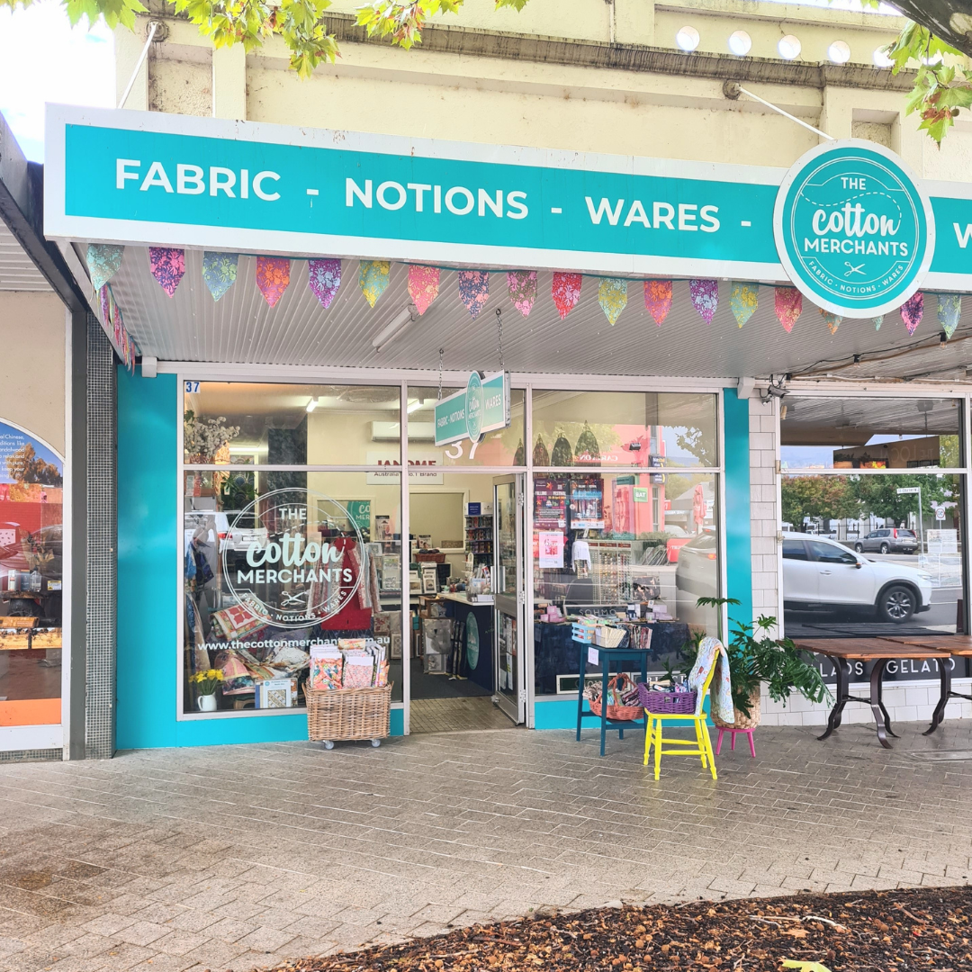 Storefront of The Cotton Merchants store with a bright blue sign advertising fabric, notions, and wares, decorated with colourful paper pennant banners outside and a display of various textile and craft items in the window.
