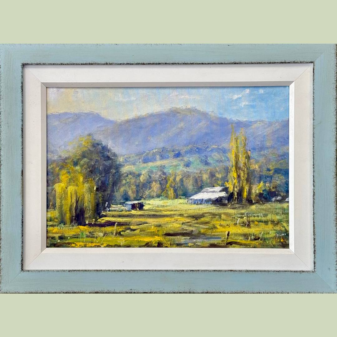 Landscape painting of a rural scene with trees, a small building, and mountains in the background, framed in a distressed light blue and white wooden frame.