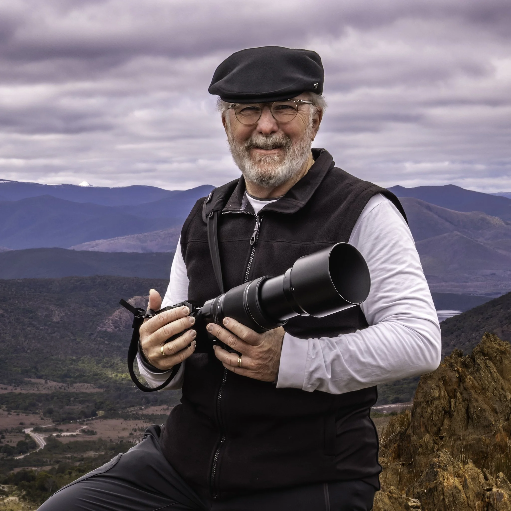 A man with glasses, a grey beard, and a black beret, smiling, holding a camera with a large telephoto lens, sitting outdoors on a rocky landscape with mountains and cloudy sky in the background.