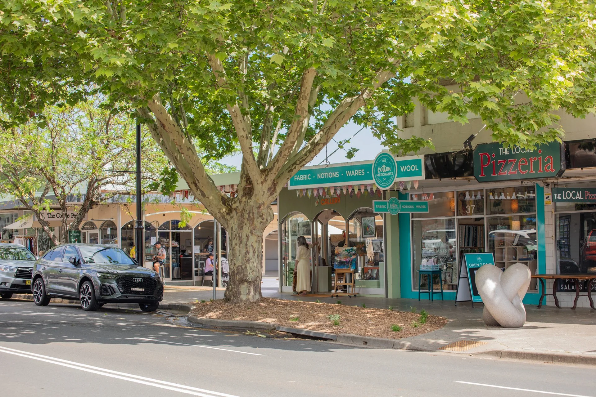 A street view with a large tree with green leaves in front of a store called The Cotton Merchants. There are cars parked on the street, and a woman in a cream dress is looking at artwork inside another shop. A sign indicates the store is open 7 days a week. A bench-shaped sculpture is on the sidewalk near the store entrance.