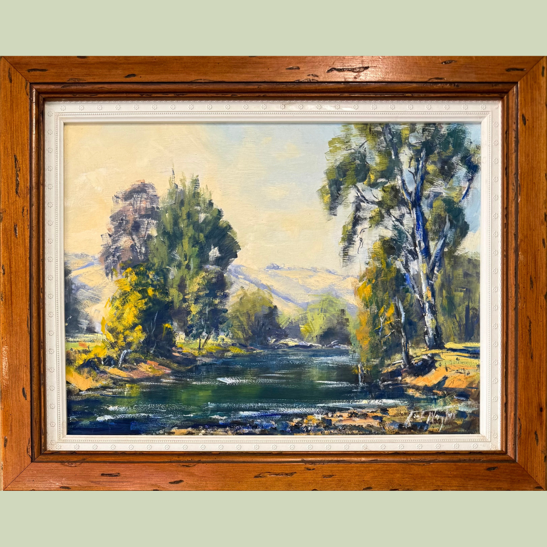 A landscape oil painting of trees and a river, with mountains and a sky in the background, framed in wood.