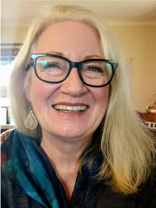 A woman smiling with long blonde hair, glasses, and teardrop earrings, indoors with a window and dining room in the background.