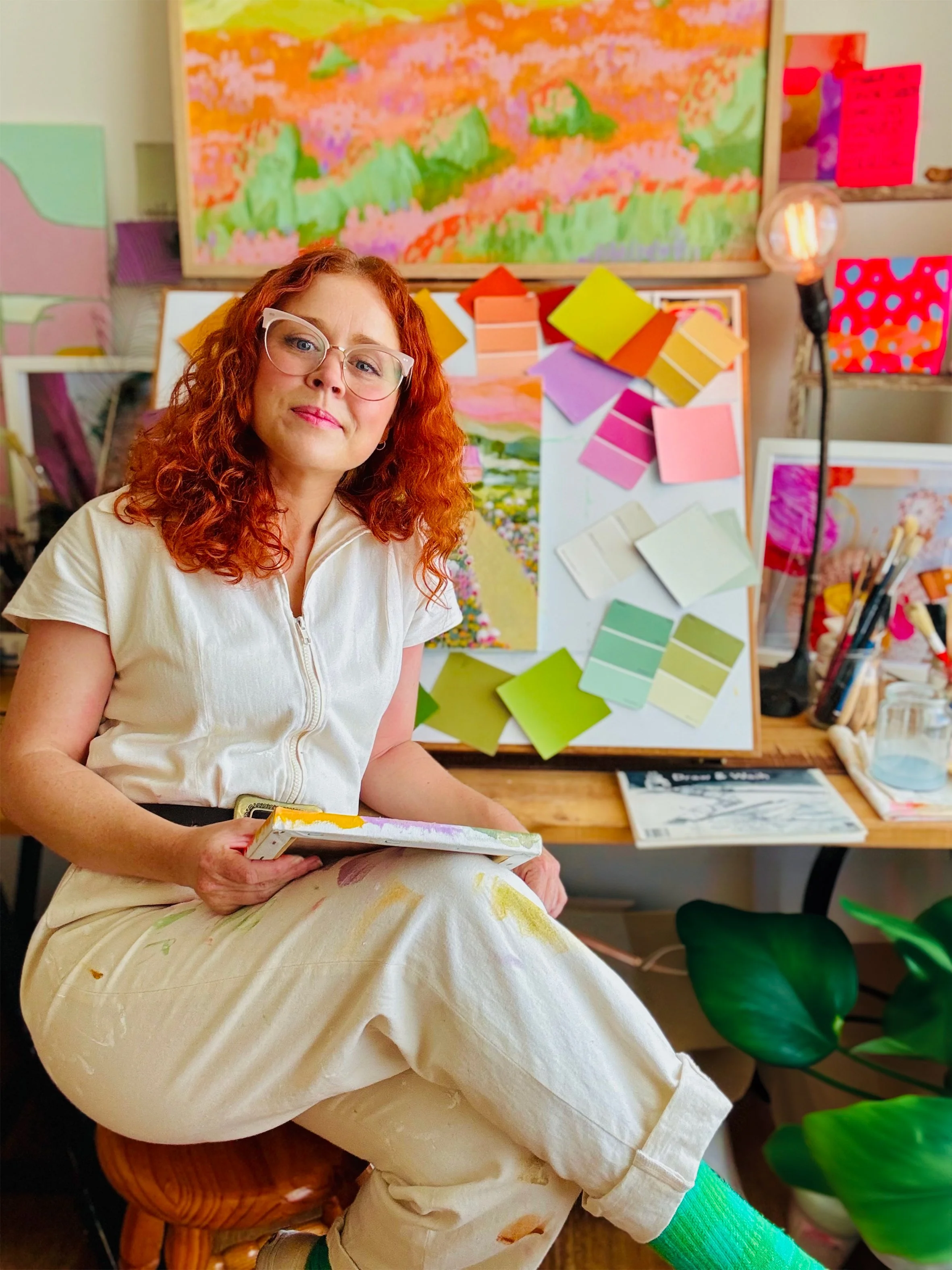 A red-haired woman with glasses sits on a stool in an art studio, holding a palette. Behind her are colorful paintings, swatches of paint, and art supplies on a table.