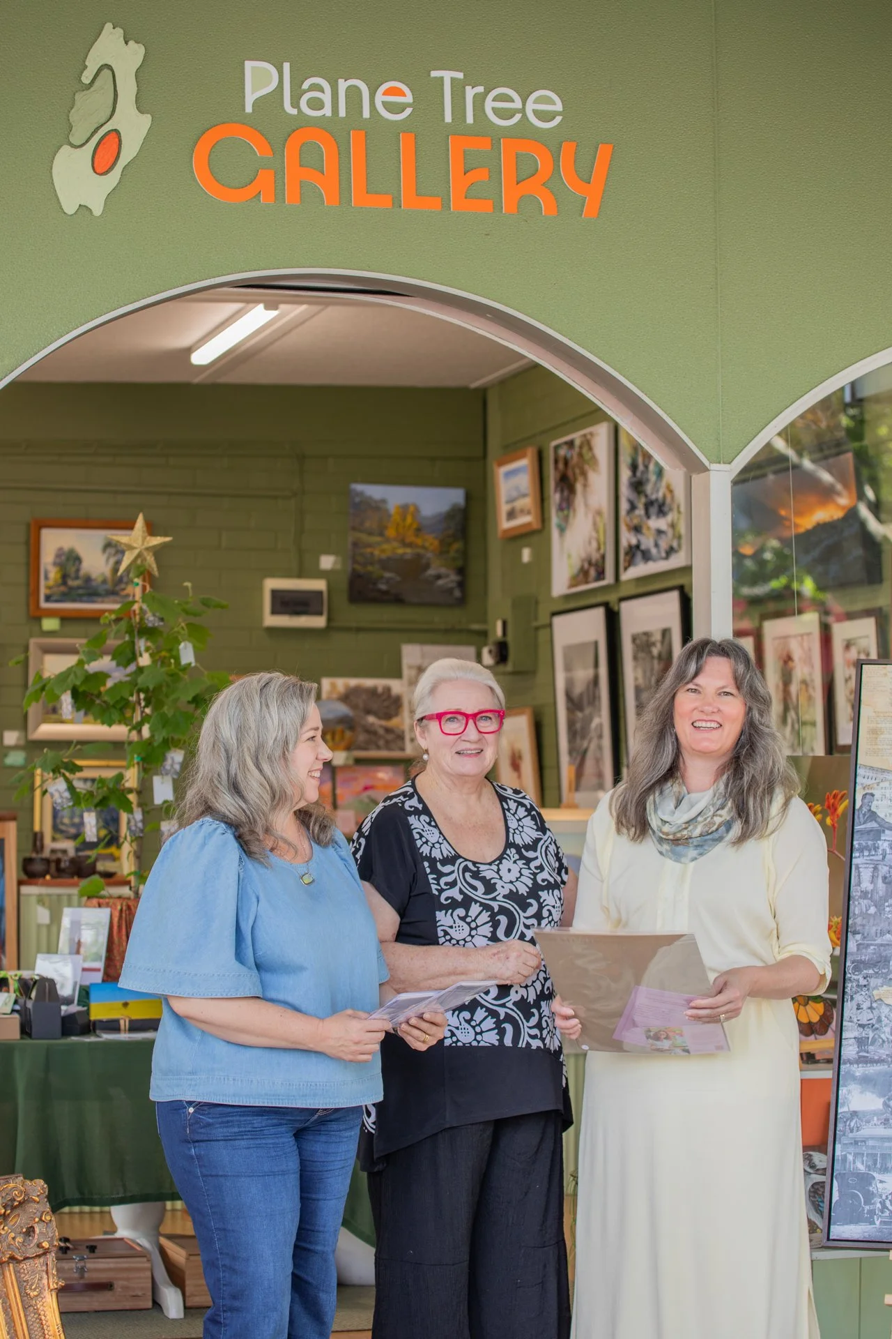 Three women standing inside the Plane Tree Gallery, surrounded by artwork on the walls and display tables, engaging in conversation and smiling.