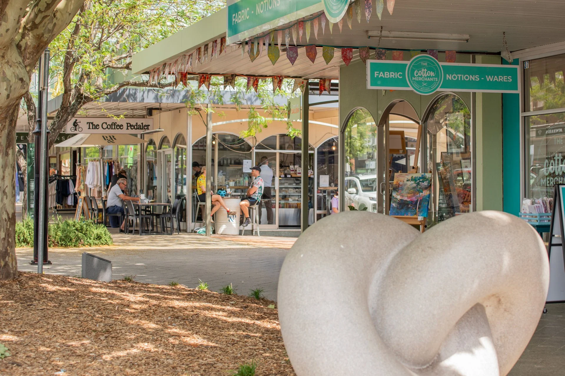 Outdoor shopping and dining area with storefronts, people sitting at tables, and a large abstract sculpture in the foreground.