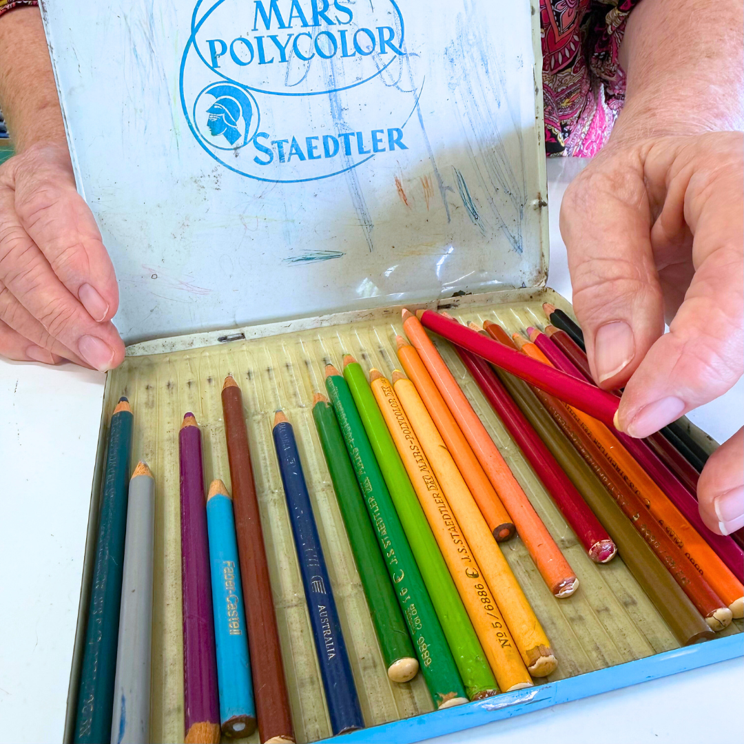 Close-up of a vintage box of colored pencils, some being used, with a person's hands opening the lid. The box has the words "Mars Polycolor" and "Staedtler" printed on it.