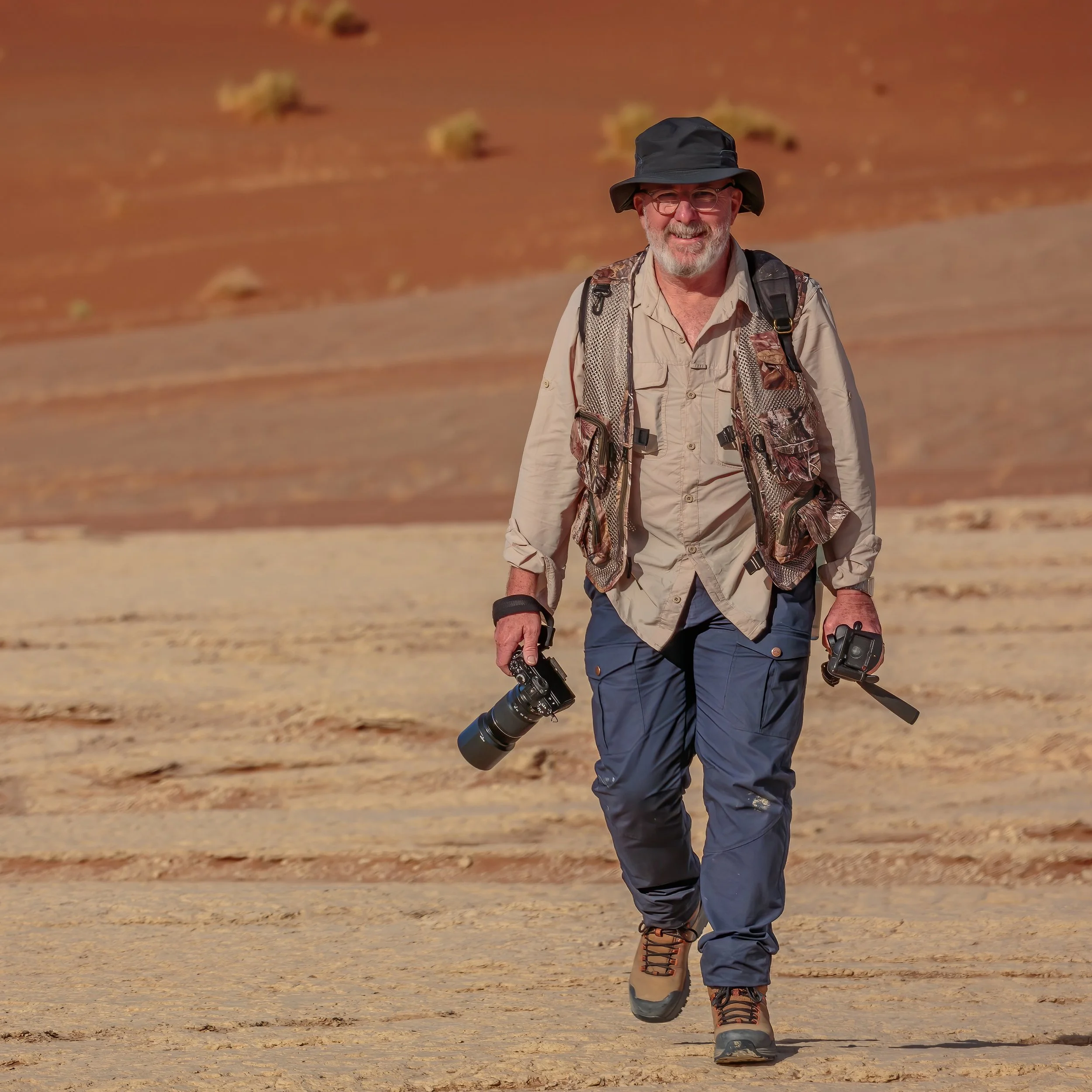 A man hiking outdoors in a desert landscape, carrying a camera and trekking poles.