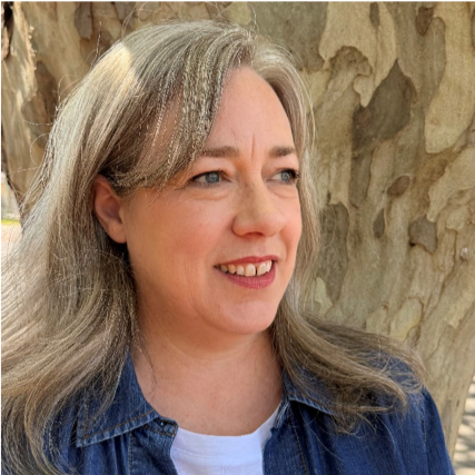 Close-up of a woman with shoulder length hair smiling outdoors against a Plane tree.