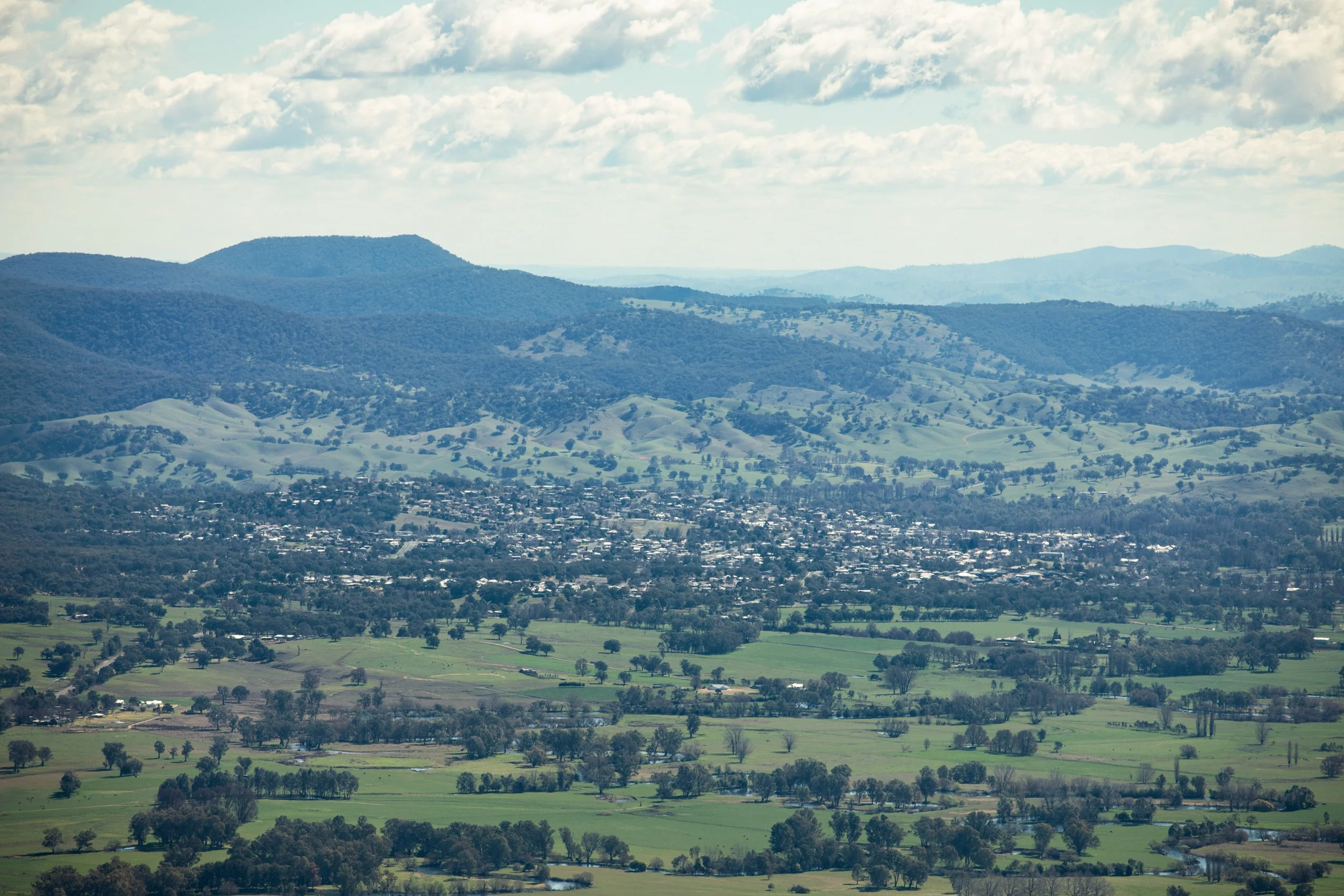 Landscape of rolling green hills and mountains with scattered trees and a small town of Tumut in the valley beneath a partly cloudy sky.