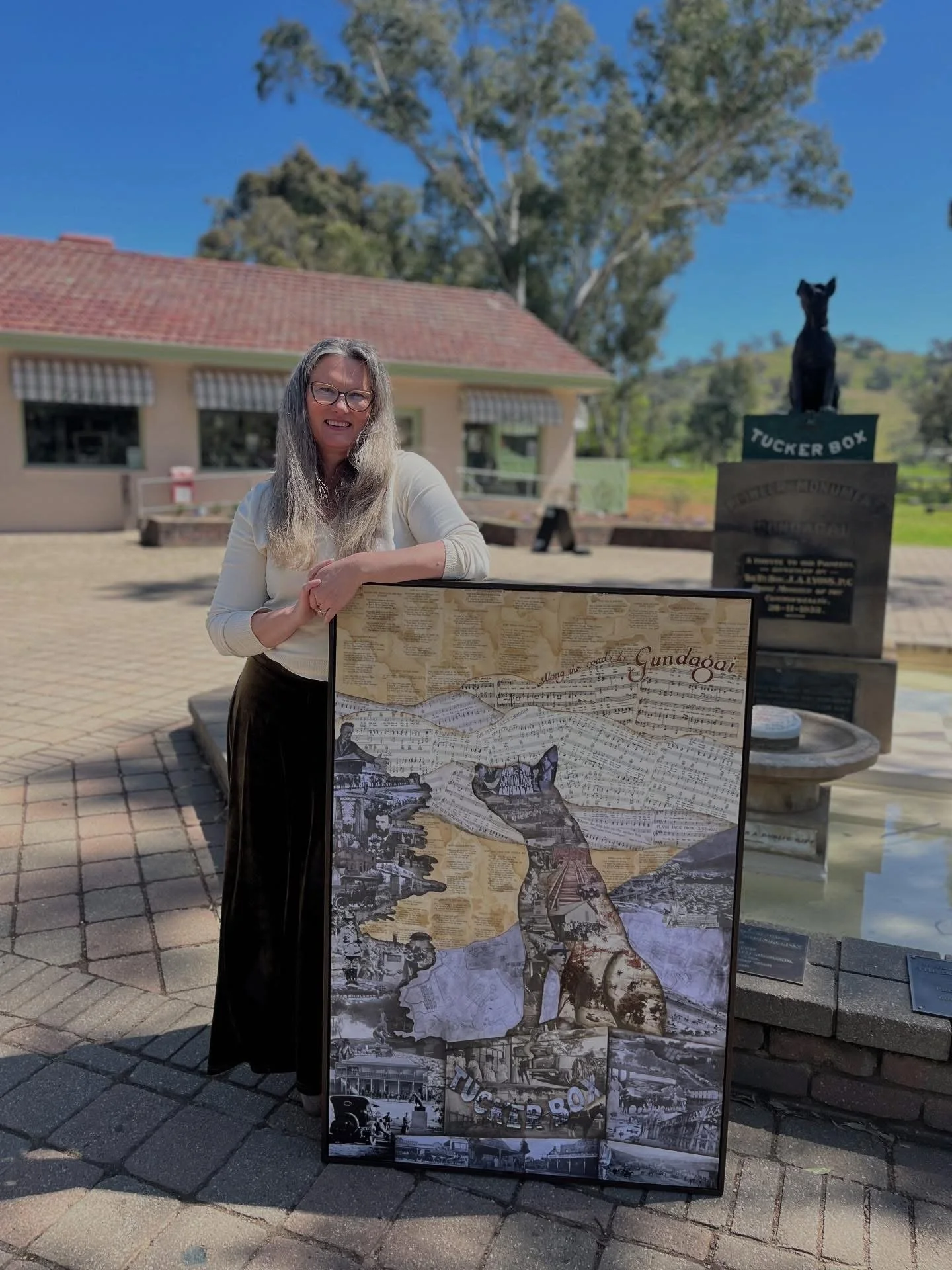 A woman with long gray hair and glasses stands outdoors on a sunny day, smiling, next to a large collage artwork. Behind her is a building with a red-tiled roof and striped awnings, and a statue of a black dog atop a pedestal. The collage features various images