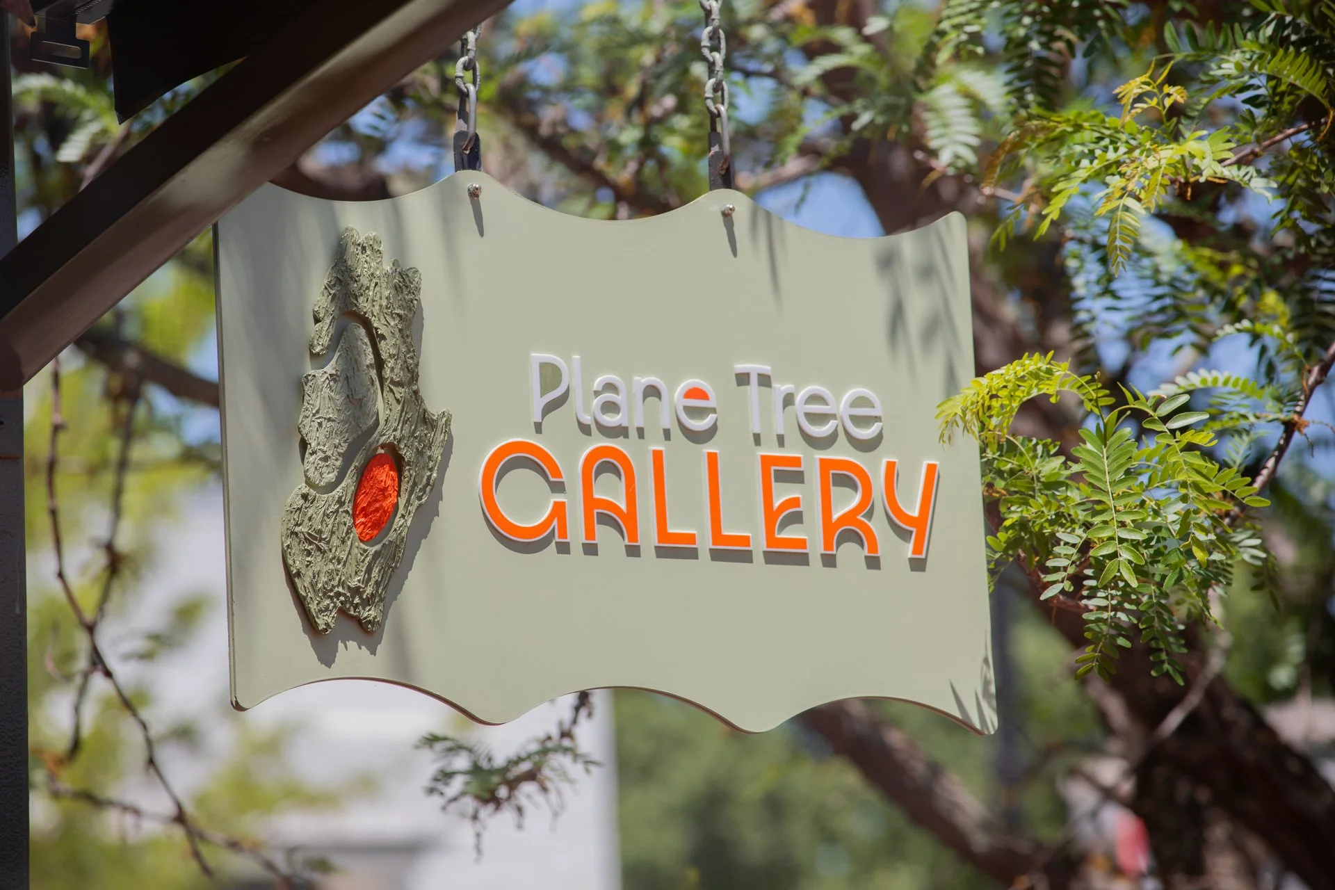 Sign for the Plane Tree Gallery with a textured tree branch and orange accents, surrounded by lush green foliage and branches, hanging outdoors.