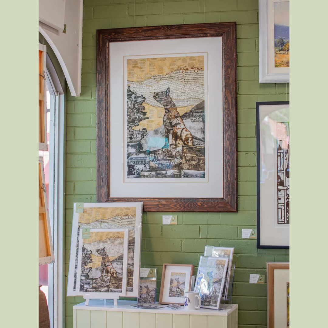 Framed artwork of a collage-style illustration featuring a dog sitting on a landscape with the word "Gundagai" at the top, displayed on a green brick wall. Several smaller framed pieces and prints of similar artwork are on a white shelf below it.