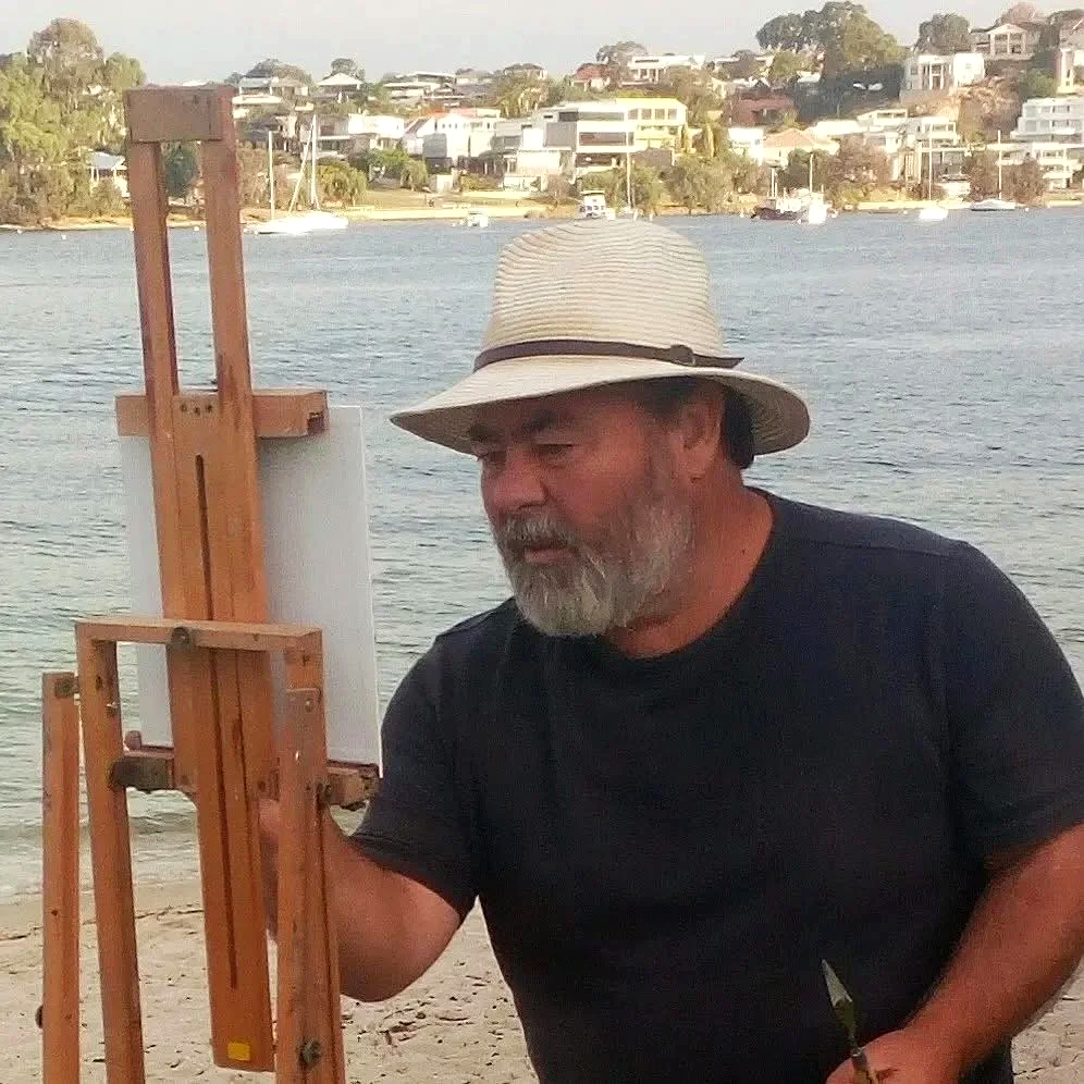 An artist painting outdoors near a body of water with boats and houses in the background.