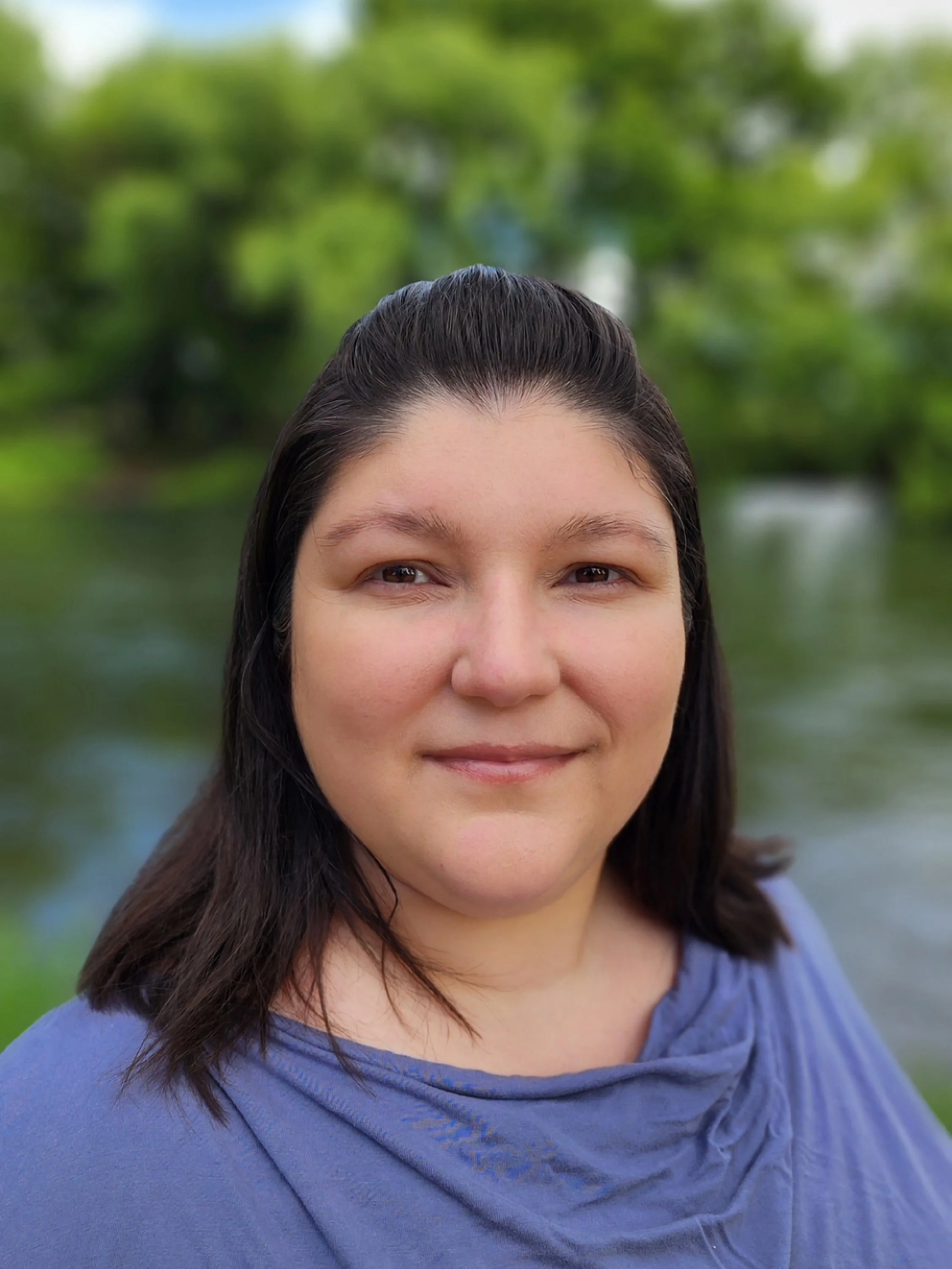 A woman with dark hair smiling outdoors near a river with green trees in the background.
