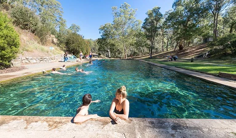 People enjoying swimming and relaxing in a large outdoor thermal pool surrounded by trees and grassy areas on a sunny day.