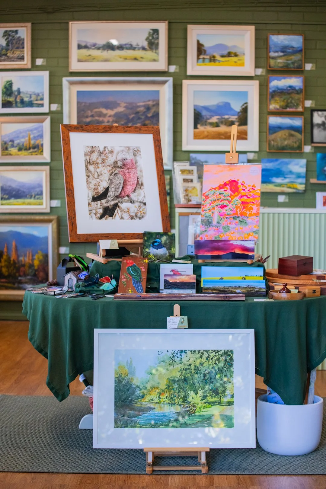 An art gallery with landscape paintings on a green wall, featuring a table with various small artworks, a framed painting of a bird, a colorful abstract painting, and a landscape painting of a river and trees in a white frame.