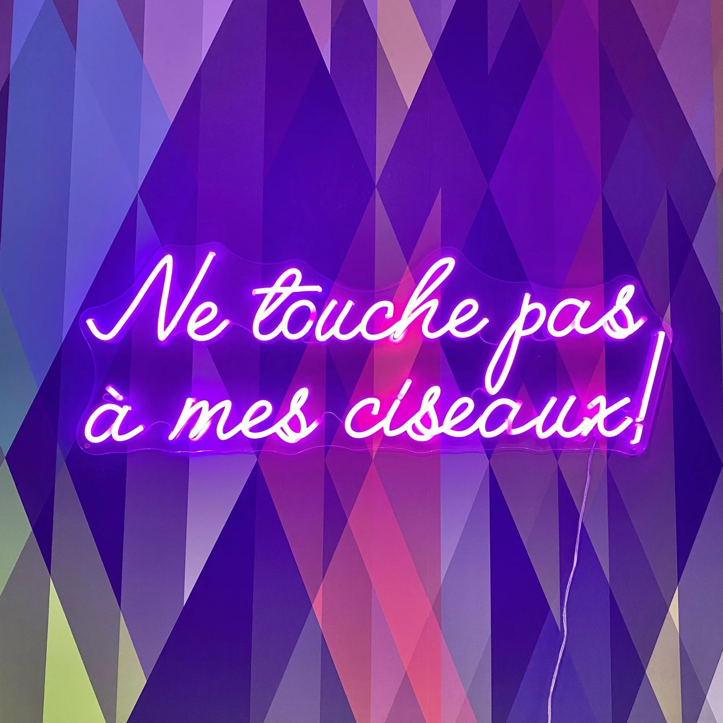 Ne touche pas à mes ciseaux! written in pink neon script against a colorful geometric patterned background.