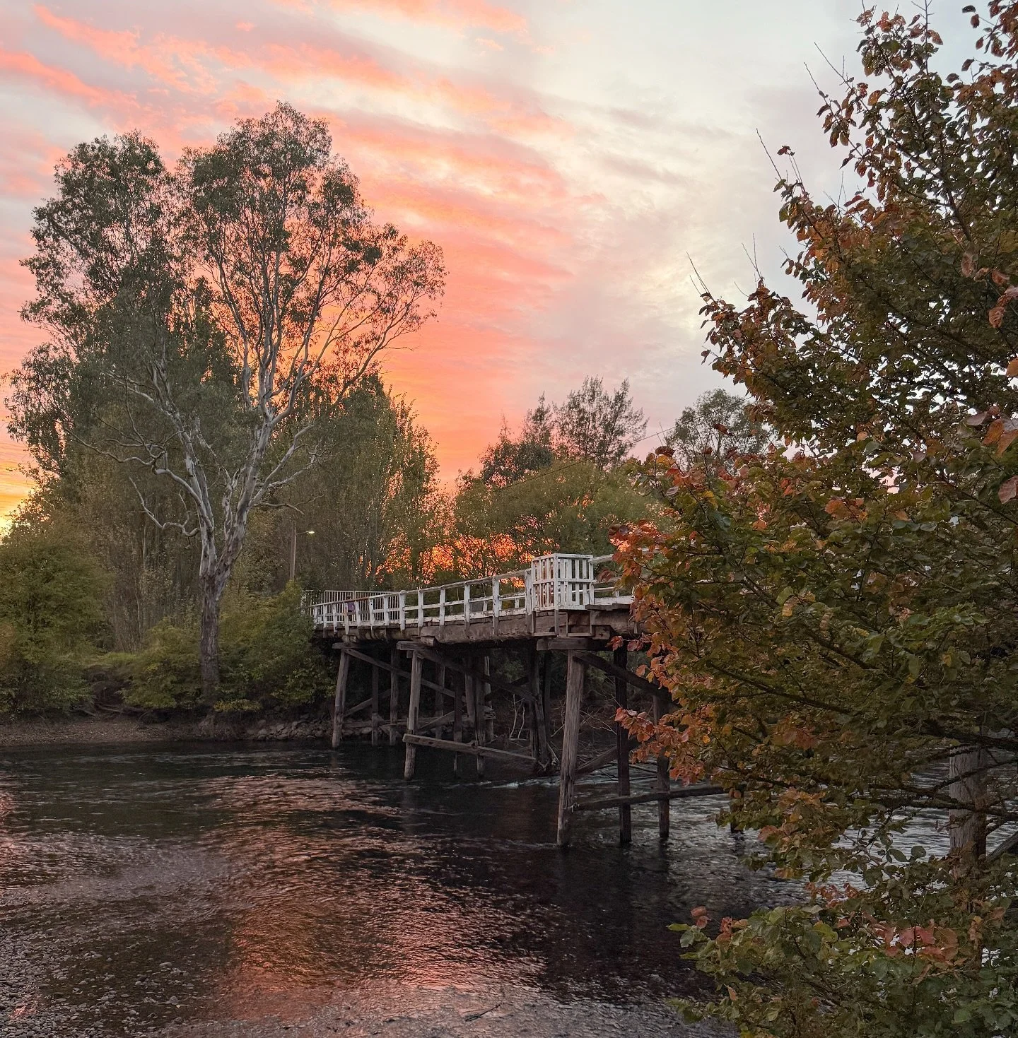 A panoramic view of a valley with lush green fields, scattered trees, and the town of Tumut in the distance, surrounded by rolling hills and mountains under a partly cloudy sky.
