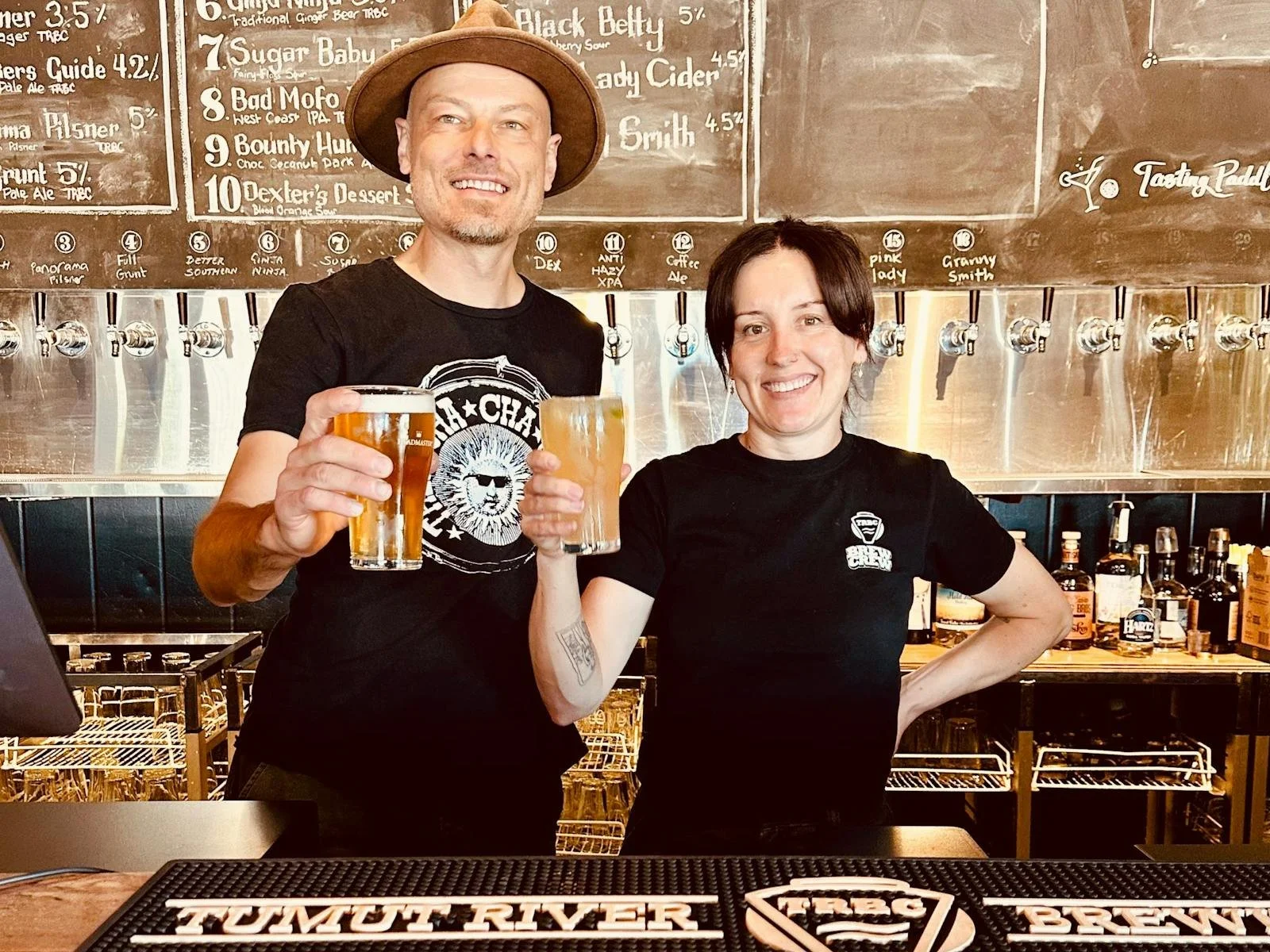 Two bartenders celebrating with drinks behind the bar at a brewery. The man is wearing a hat and a black T-shirt with a white logo, and the woman is smiling and wearing a black brewery T-shirt. The background shows a chalkboard menu and beer taps.