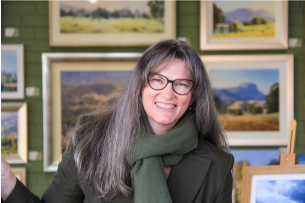Woman with long hair, glasses, and green scarf smiling, standing in front of landscape paintings in an art gallery.