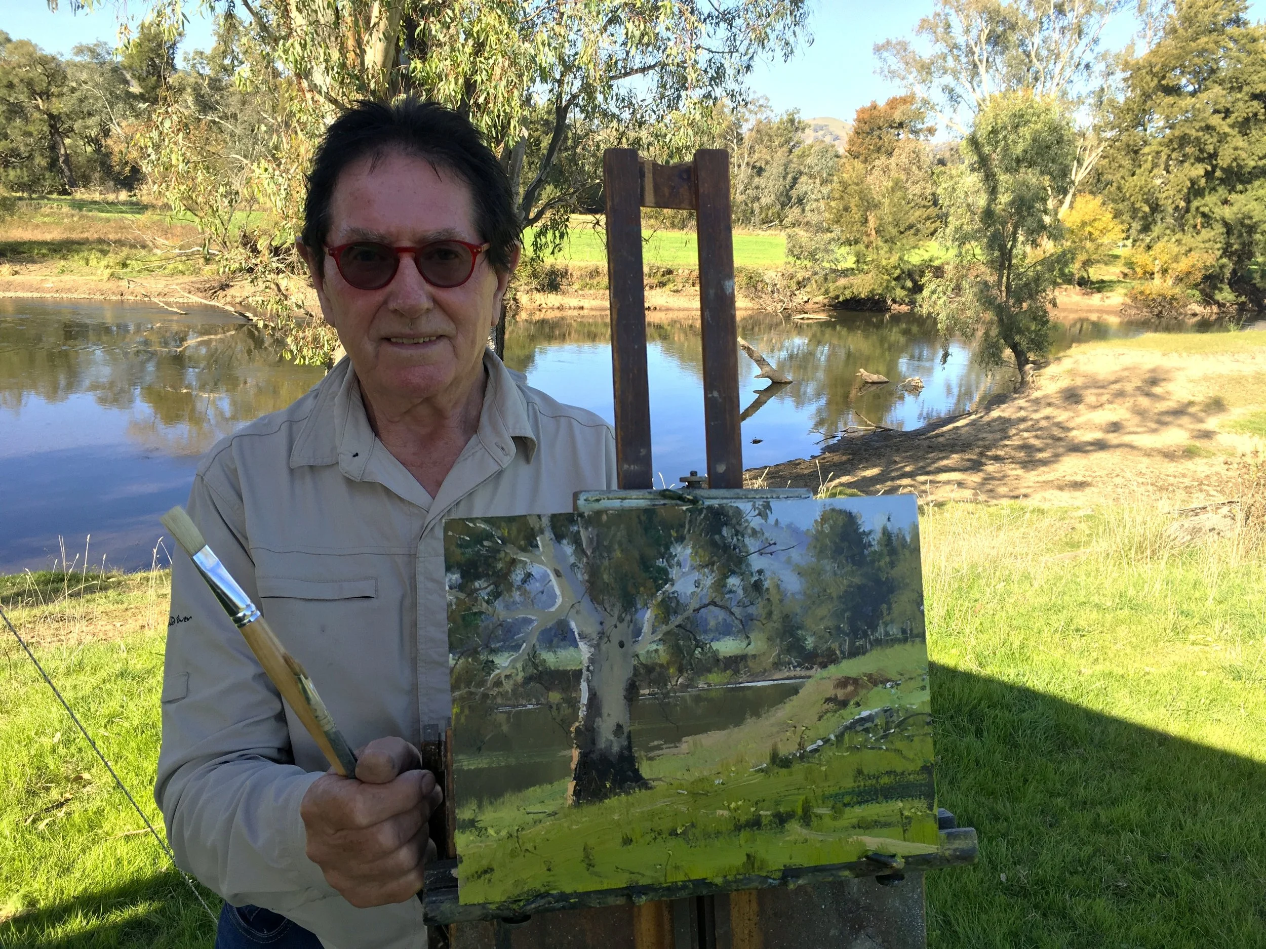 Person with red glasses and beige shirt outdoors near a river, holding a paintbrush and displaying a painting of a tree and water scene.