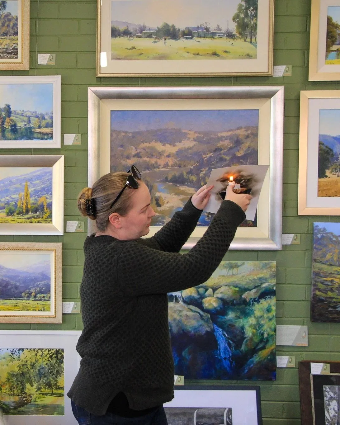 A woman in a black sweater with sunglasses on her head holding a photograph with a lit matchstick towards the image. She is standing in front of a green brick wall decorated with various landscape paintings.